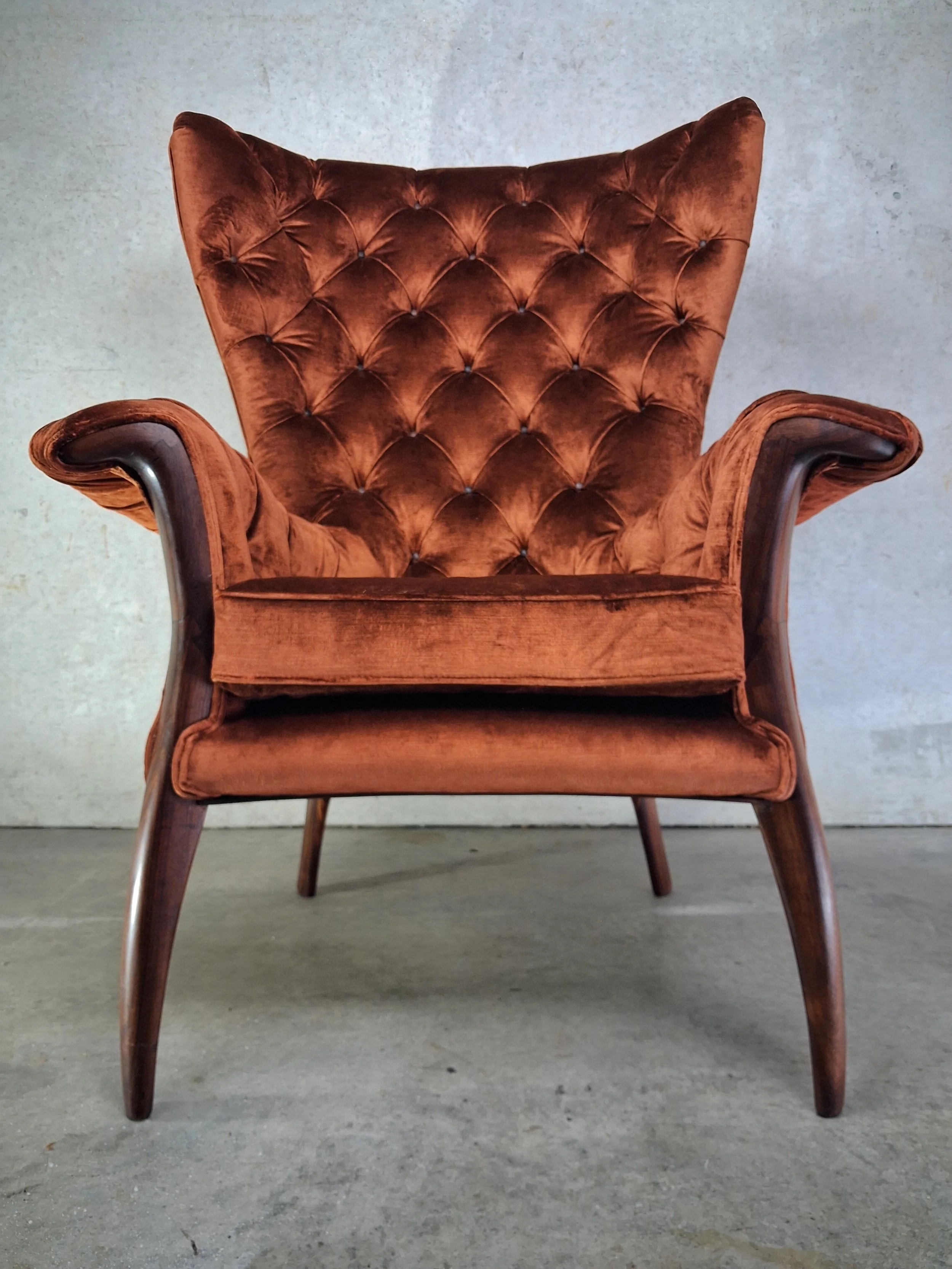 A custom-made brown velvet armchair with wooden legs and a tufted backrest, placed against a concrete wall.