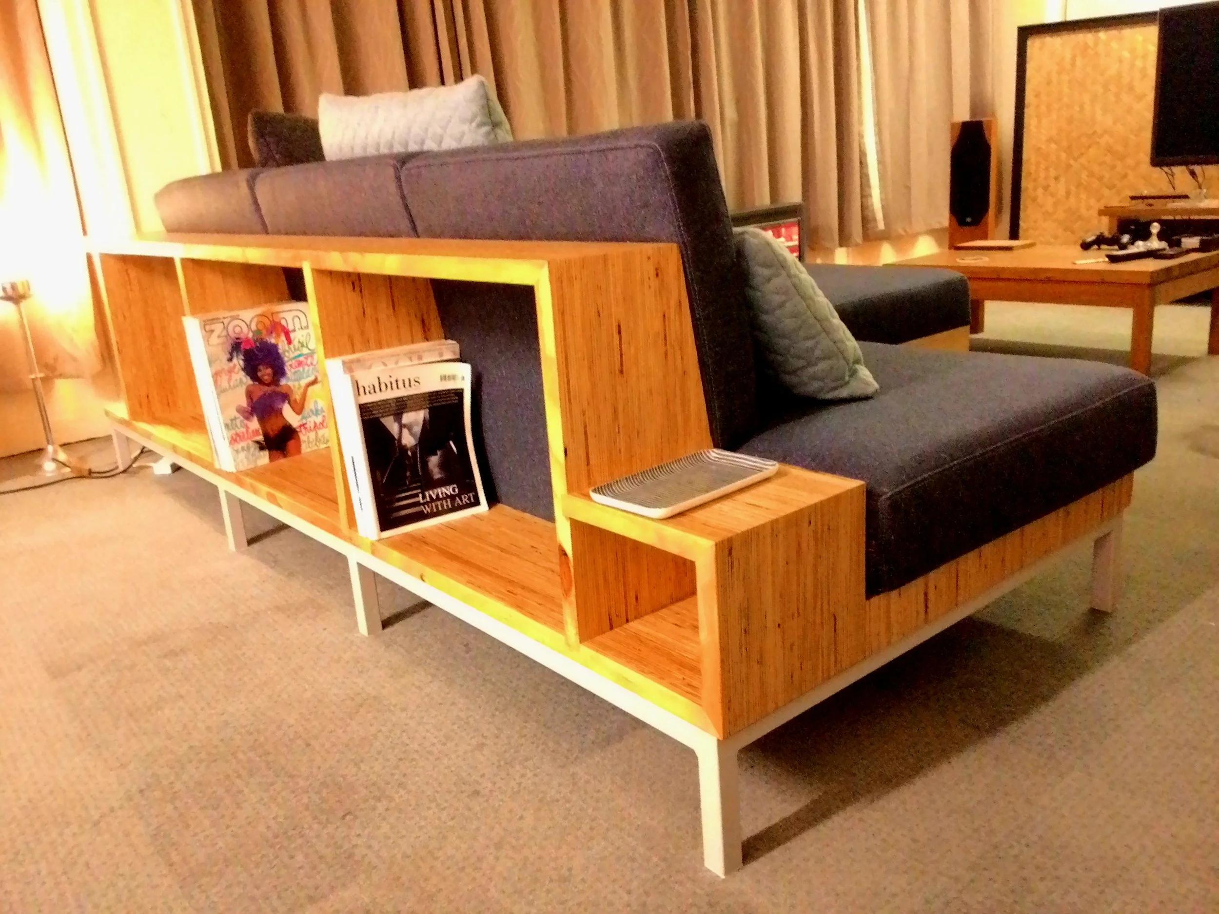 Living room with a modern wooden bookshelf and a dark upholstered sofa, books arranged inside the shelf, a heater on top, and cushions on the sofa. In the background, there are curtains, a TV, and a coffee table with remote controls.
