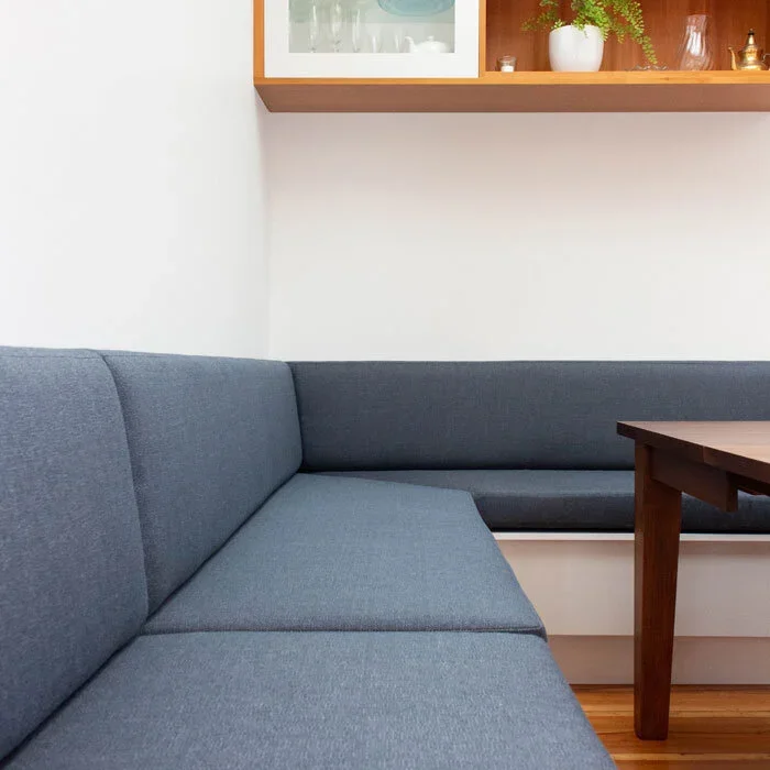 A corner of a room with a blue sectional sofa against a white wall, and a wooden shelf with decorative items and a potted plant above the sofa.