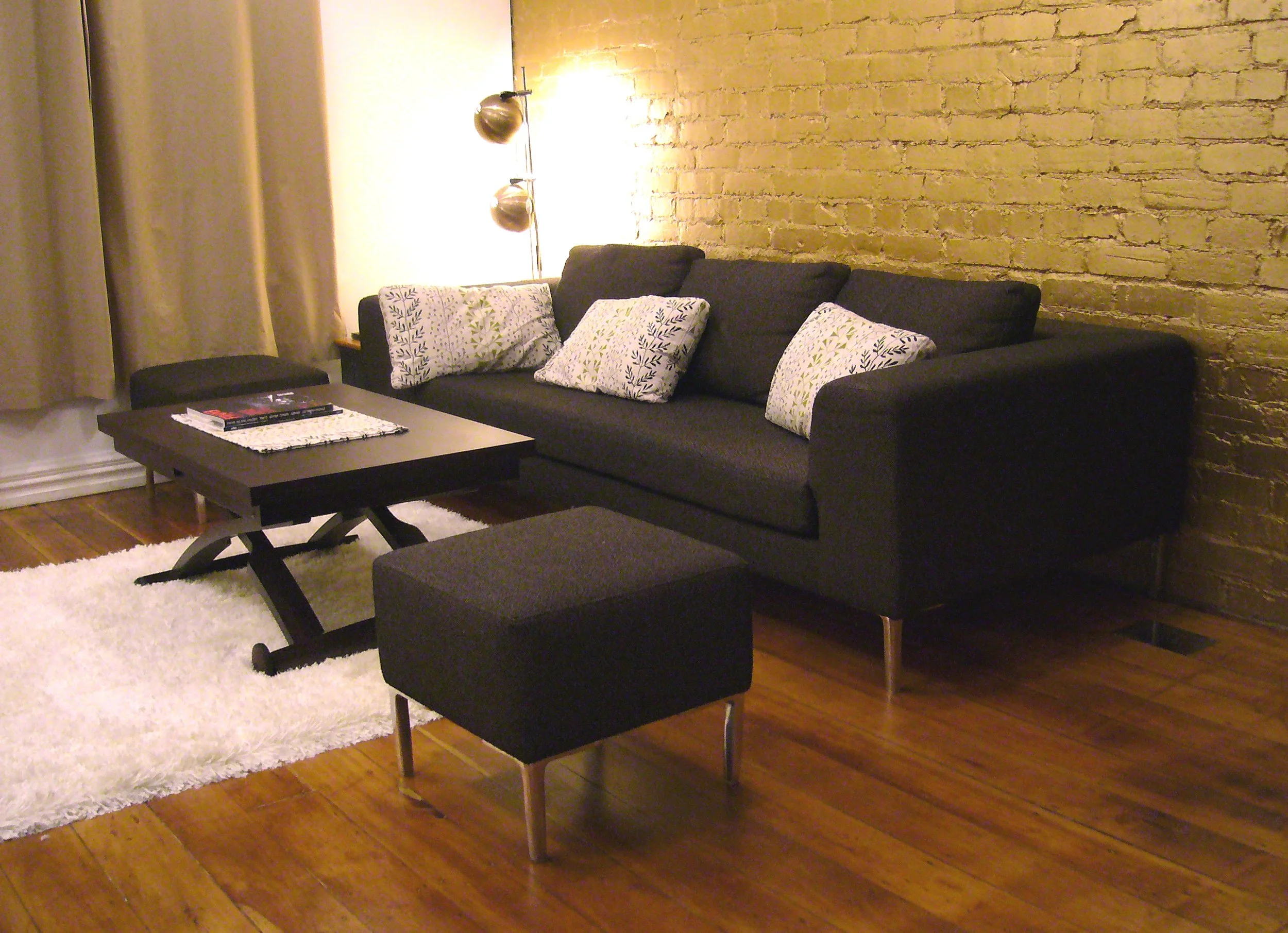 Living room with a black sofa, decorative pillows, a small black ottoman, a dark coffee table with magazines, a white fluffy rug, a brick accent wall, and a wall sconce with three bulbs.