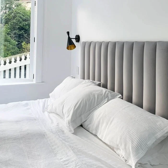 A cozy bedroom with a white bed and pillows, a large window showing greenery outside, and a black wall-mounted reading lamp.