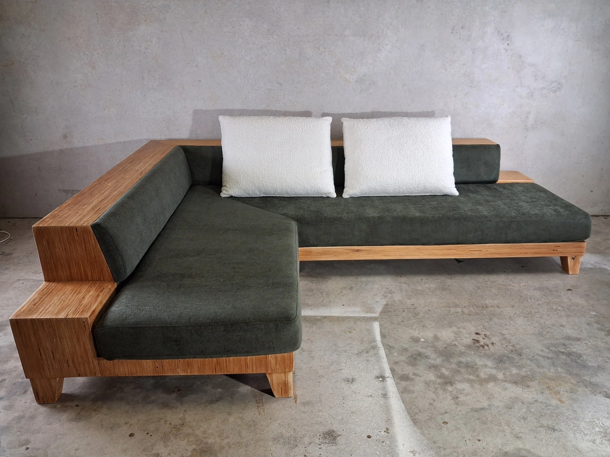 Modern green sectional sofa with wooden frame and two white pillows, set against a concrete wall and floor.