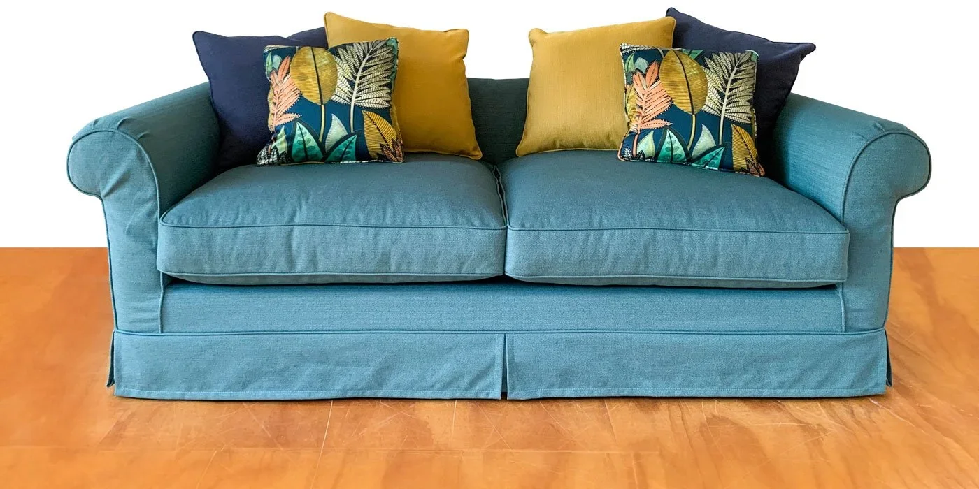 A teal sofa with rolled arms and a skirted base, adorned with four pillows: two navy ones, two yellow ones, and two botanical print pillows with leaves and ferns.
