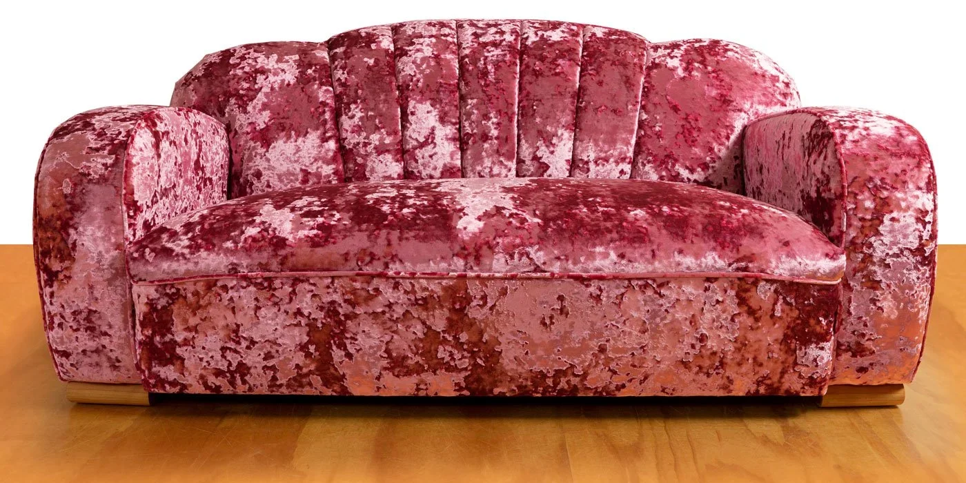 A vintage pink velvet upholstered sofa with carved wooden feet on a wooden floor.