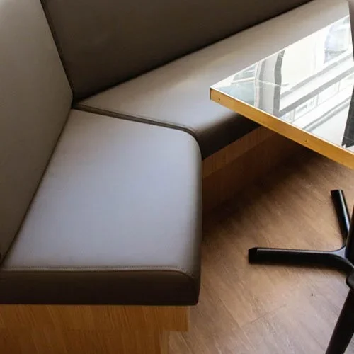 A gray upholstered booth seat with a wooden bottom, next to a glass-top table with a wooden edge, on a wooden floor.