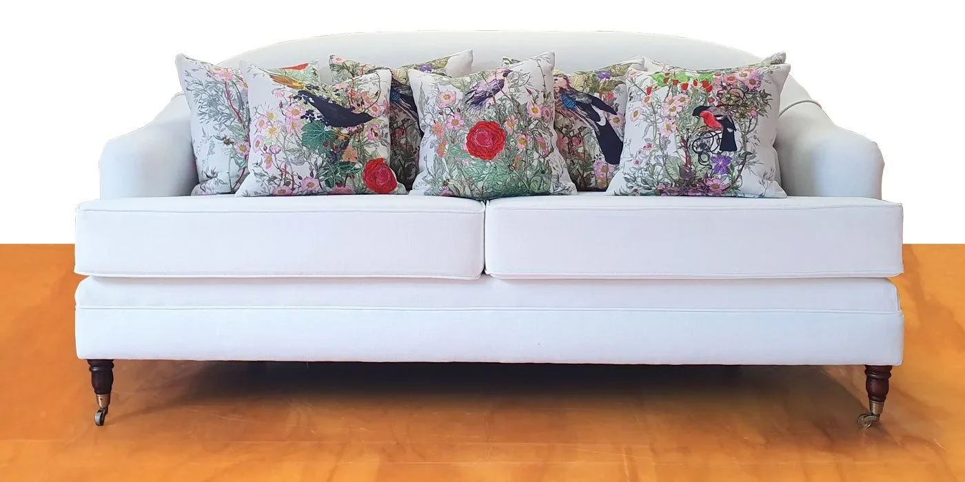 A white sofa with wooden legs and casters, decorated with multiple pillows featuring floral and bird designs.