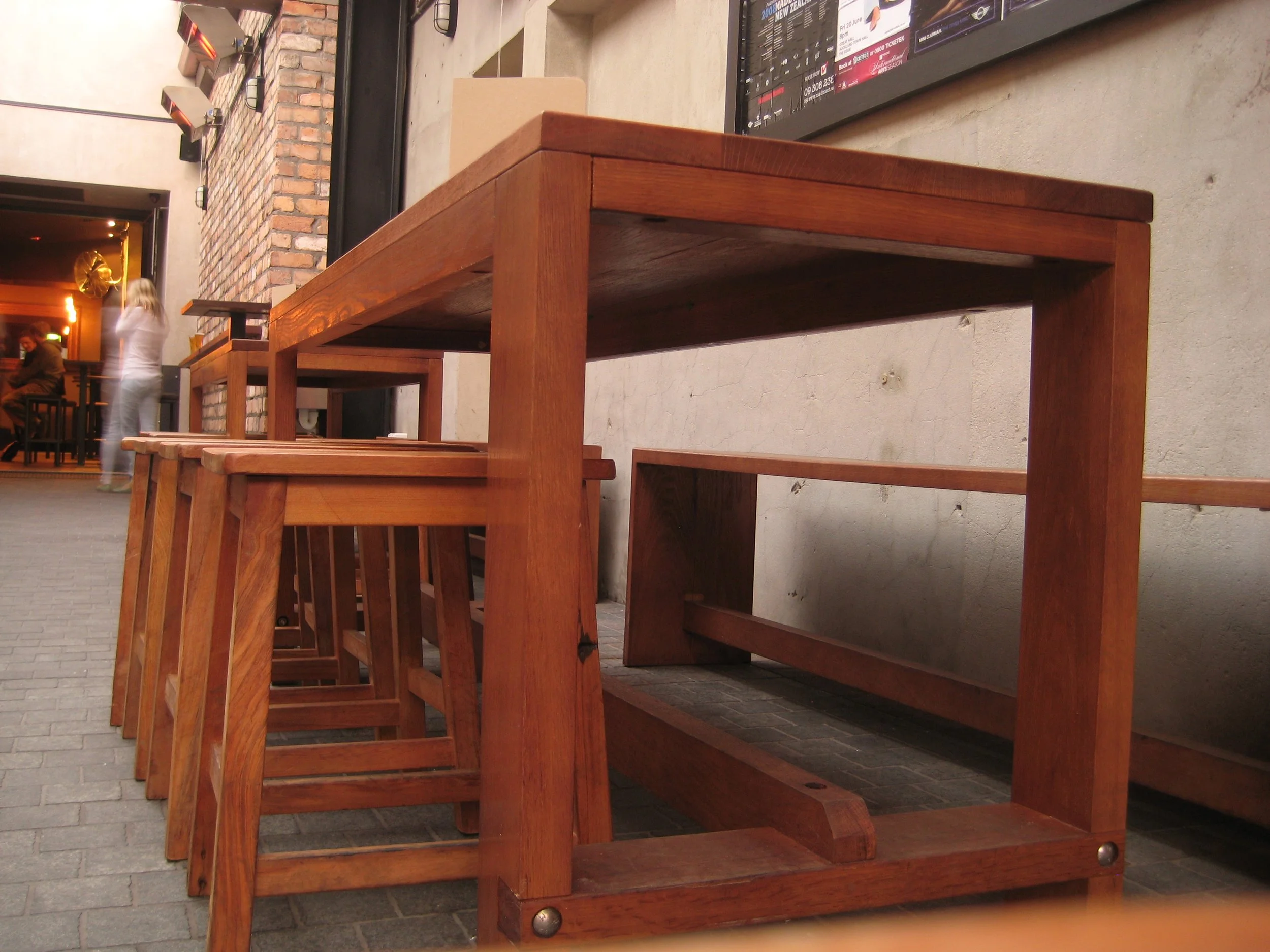 A row of wooden tables and benches outside a restaurant or cafe on a city sidewalk, with a woman and a man in the background near the entrance.