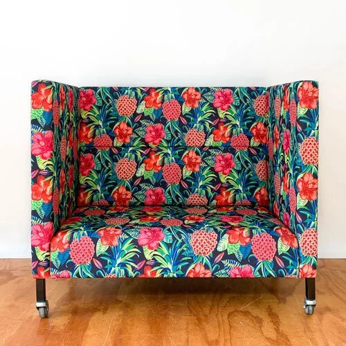 A small upholstered sofa with a colorful floral pattern featuring red, pink, and green flowers on a blue background, placed against a white wall and on a wooden floor.