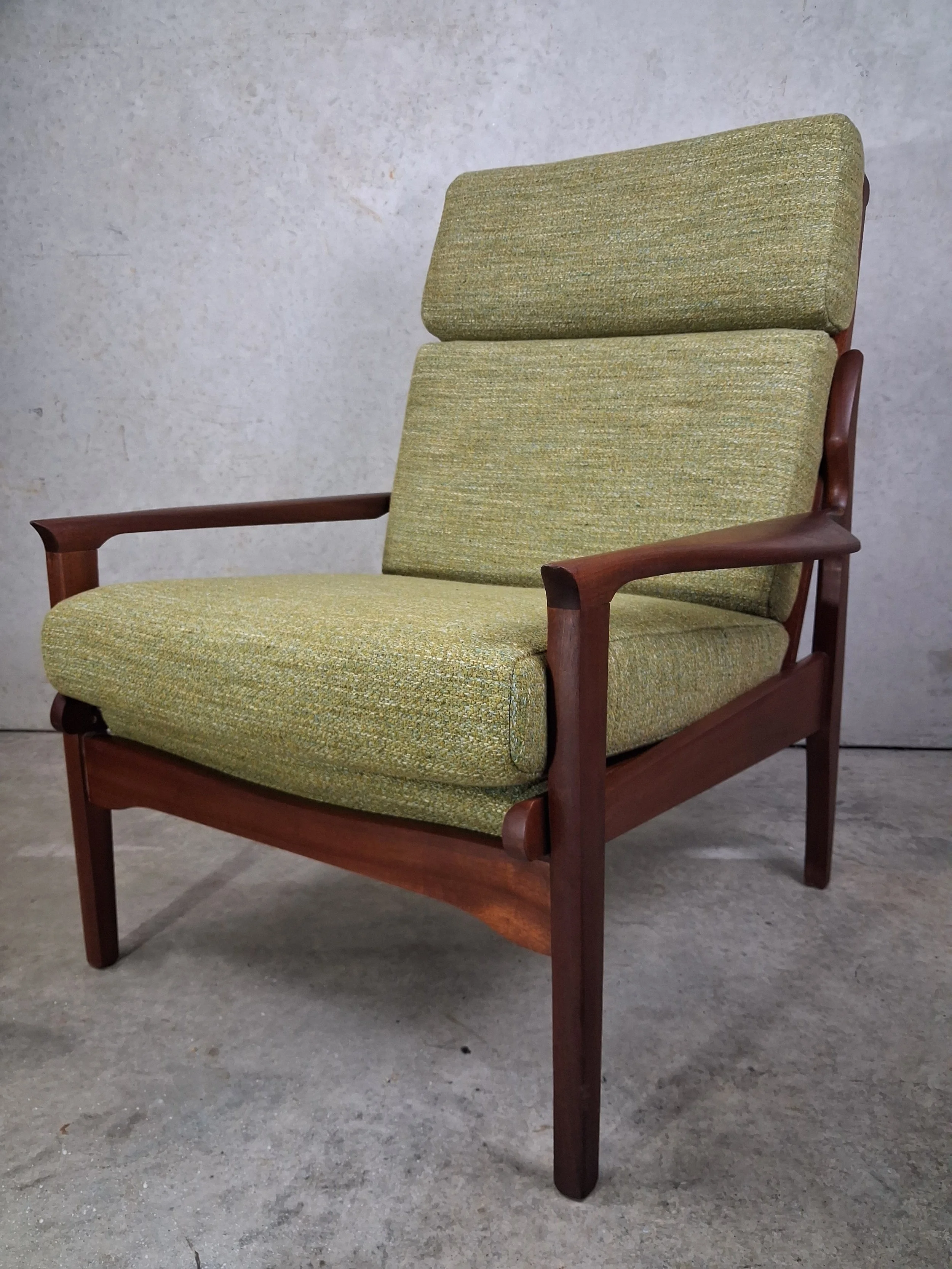 A vintage mid-century modern armchair with wooden frame and green textured fabric upholstery on the seat, backrest, and adjustable headrest, situated on a concrete floor against a plain wall.