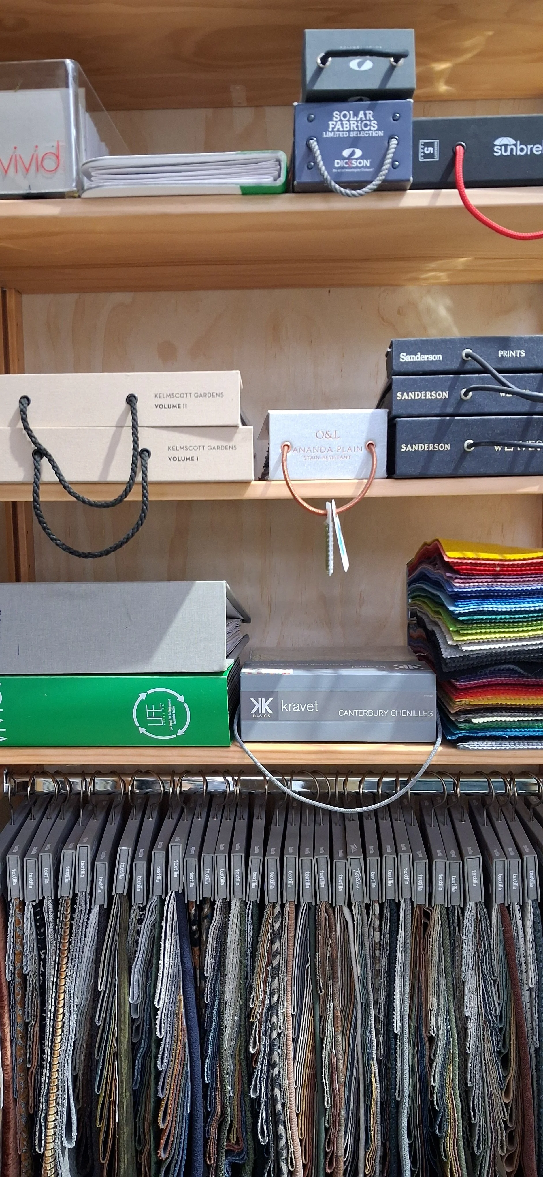 Wooden shelf displaying fabric samples, catalogs, and boxes, with fabric swatches hanging below on metal hooks.