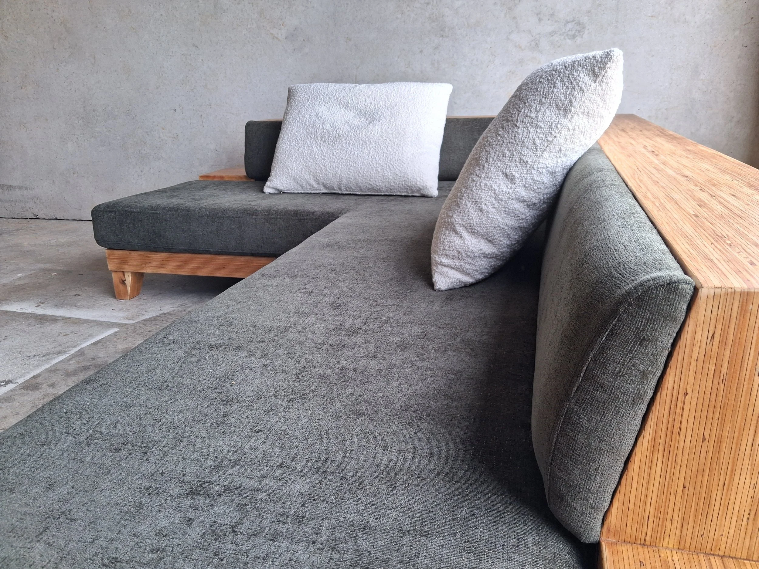 Modern grey sectional sofa with textured pillows and wooden accents, set against a concrete wall.