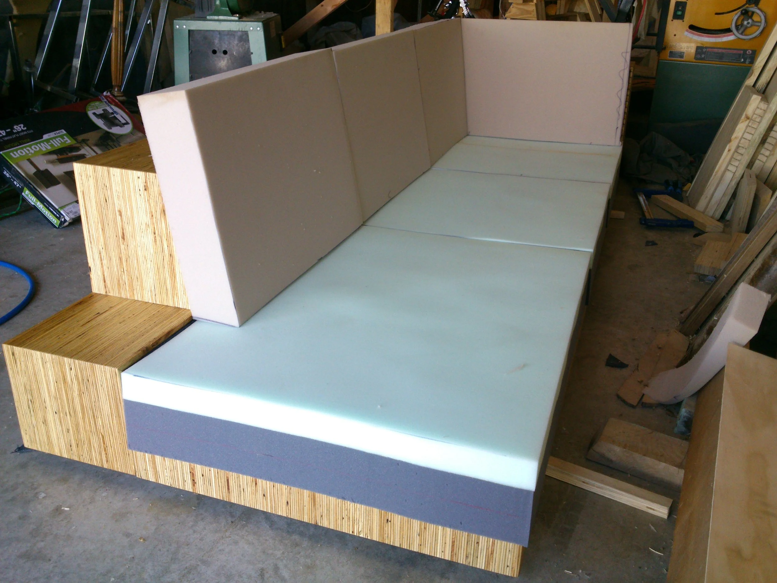 A partially assembled sofa with wooden sides, upholstered back and seat cushions, in a workshop with tools and wood scattered around.