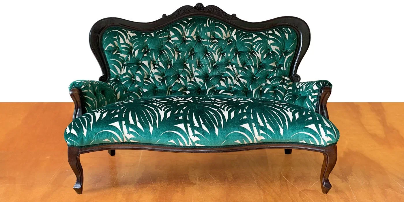 Vintage green upholstered sofa with dark wood frame and carved details on the top edge, placed on a wooden floor against a white wall.