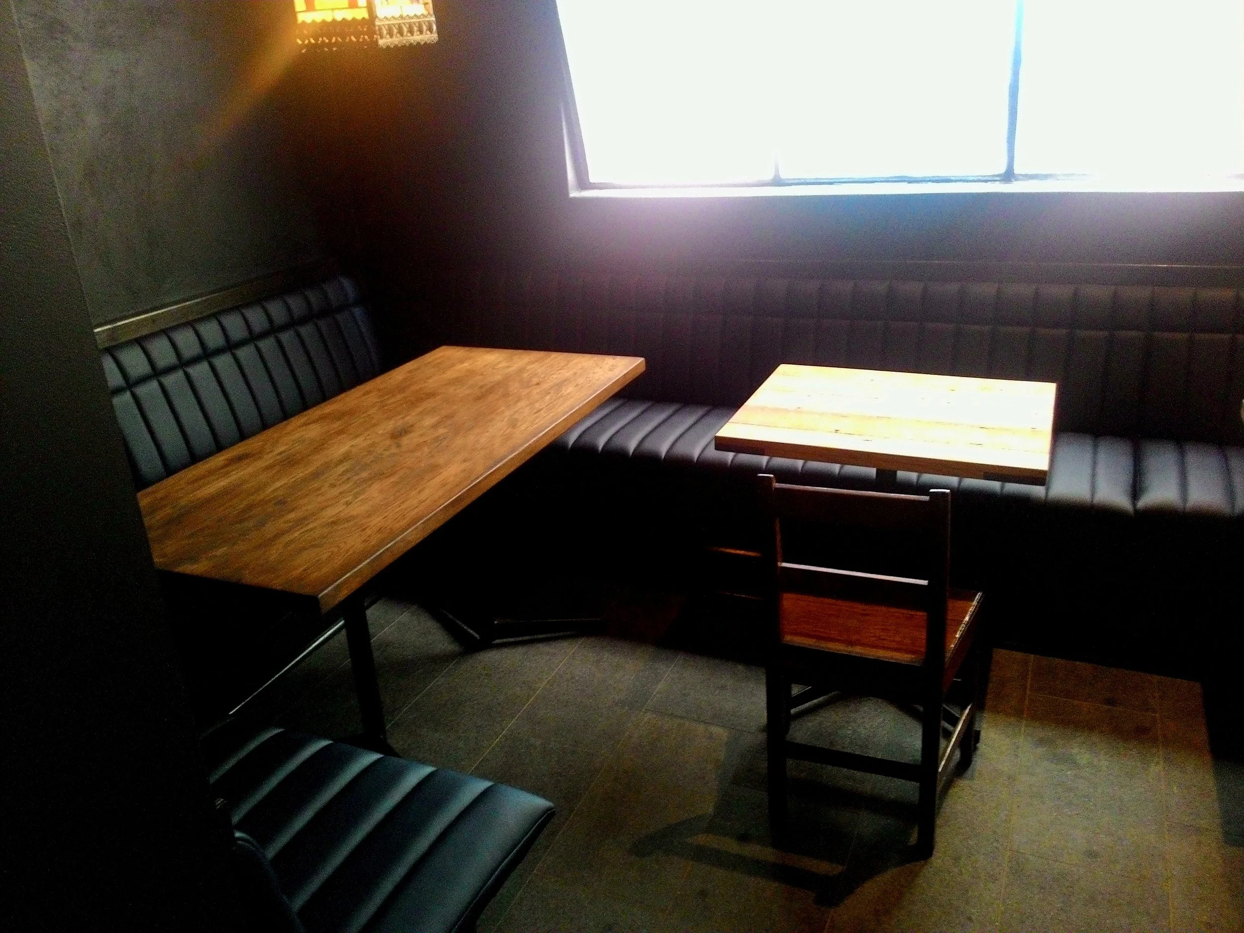 Empty restaurant corner with two wooden tables, black cushioned bench seating along the wall, a black chair, and a window allowing bright sunlight inside.