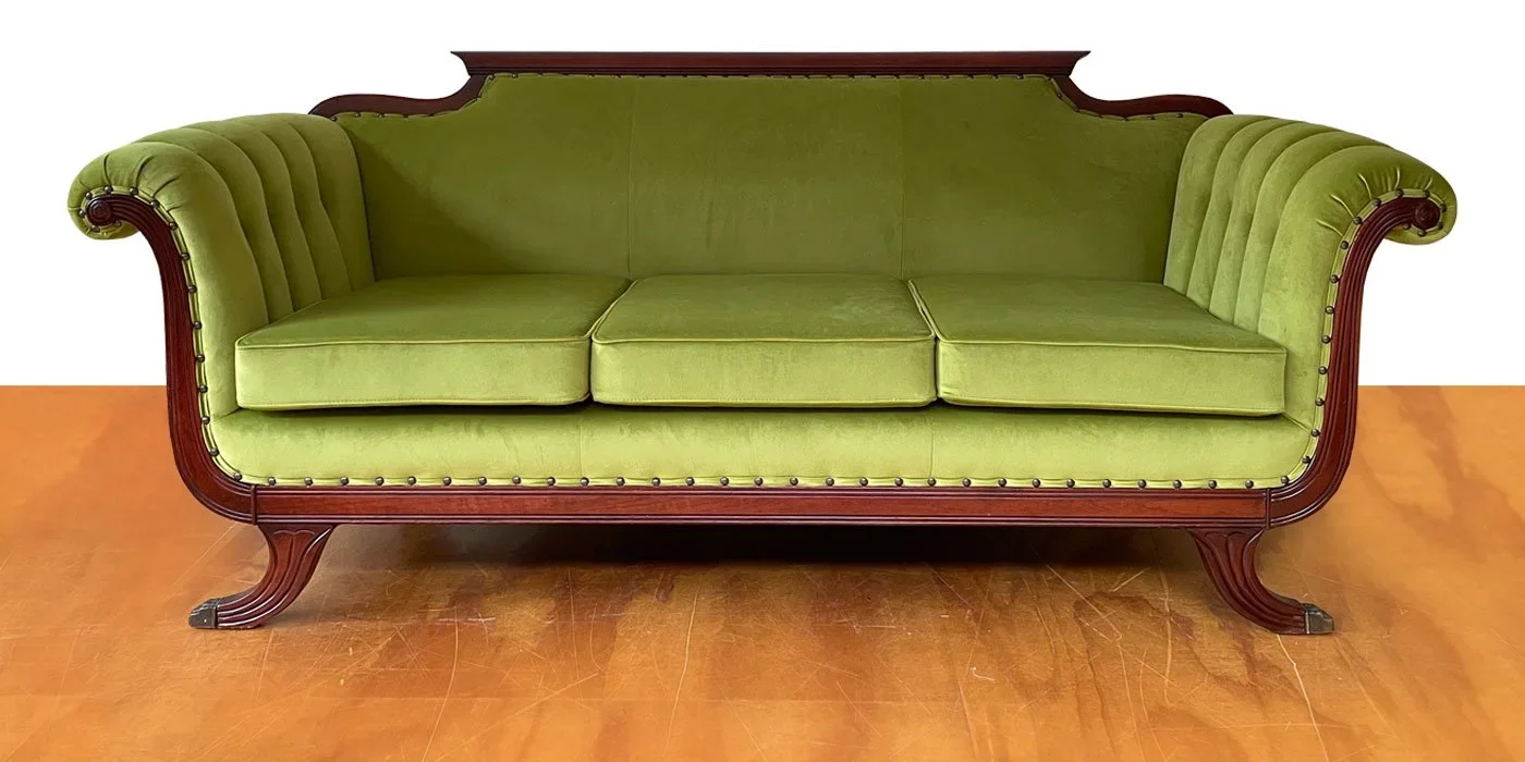 A vintage green velvet sofa with carved wood frame and nailhead trim, placed on a wooden floor.