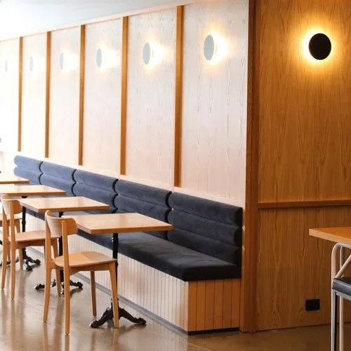 A modern restaurant interior with a row of small wooden tables and chairs, dark blue cushioned bench seating, and wooden wall paneling with circular light fixtures.