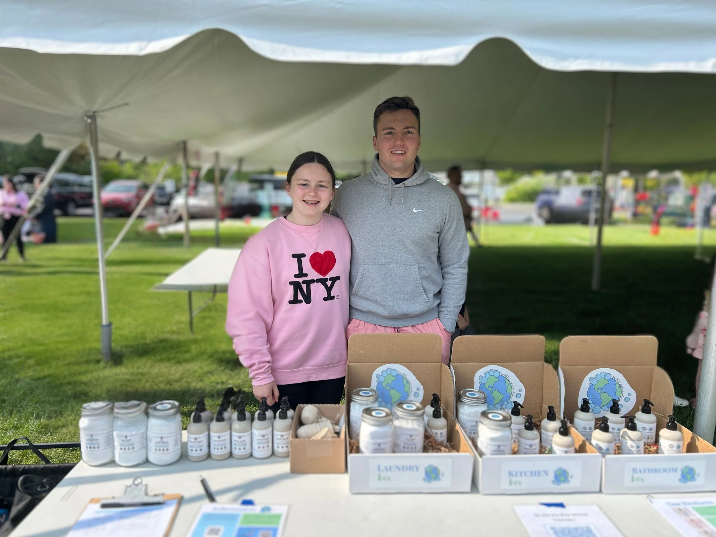A girl and a boy standing behind a table with environmentally friendly bottles and containers at an outdoor event under a large tent. There are cars and people in the background.
