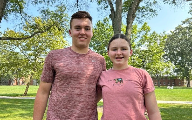 A young man and woman standing outdoors in a park with trees and grass, smiling at the camera.