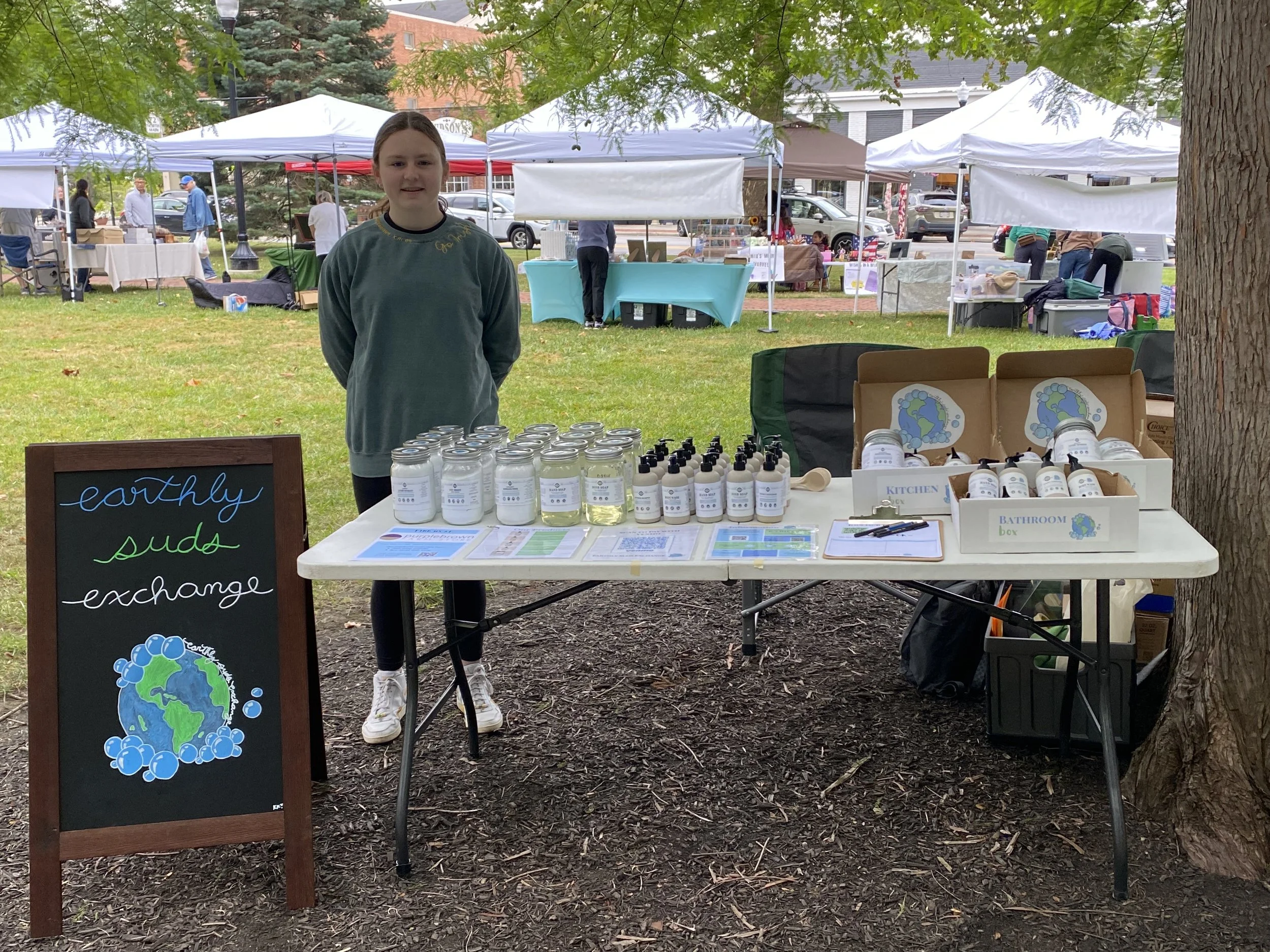 A young girl standing behind a booth at an outdoor market or fair, selling eco-friendly and earth-conscious products such as hand soap. The booth features a sign with a globe and bubble graphics, and the background shows other market stalls, trees, and people.