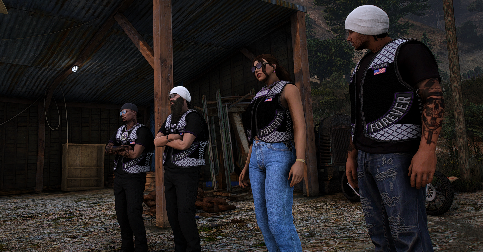 Four people standing outdoors in front of a wooden shed, wearing matching black vests with patches, some with tattoos and sunglasses, among trees and hills.