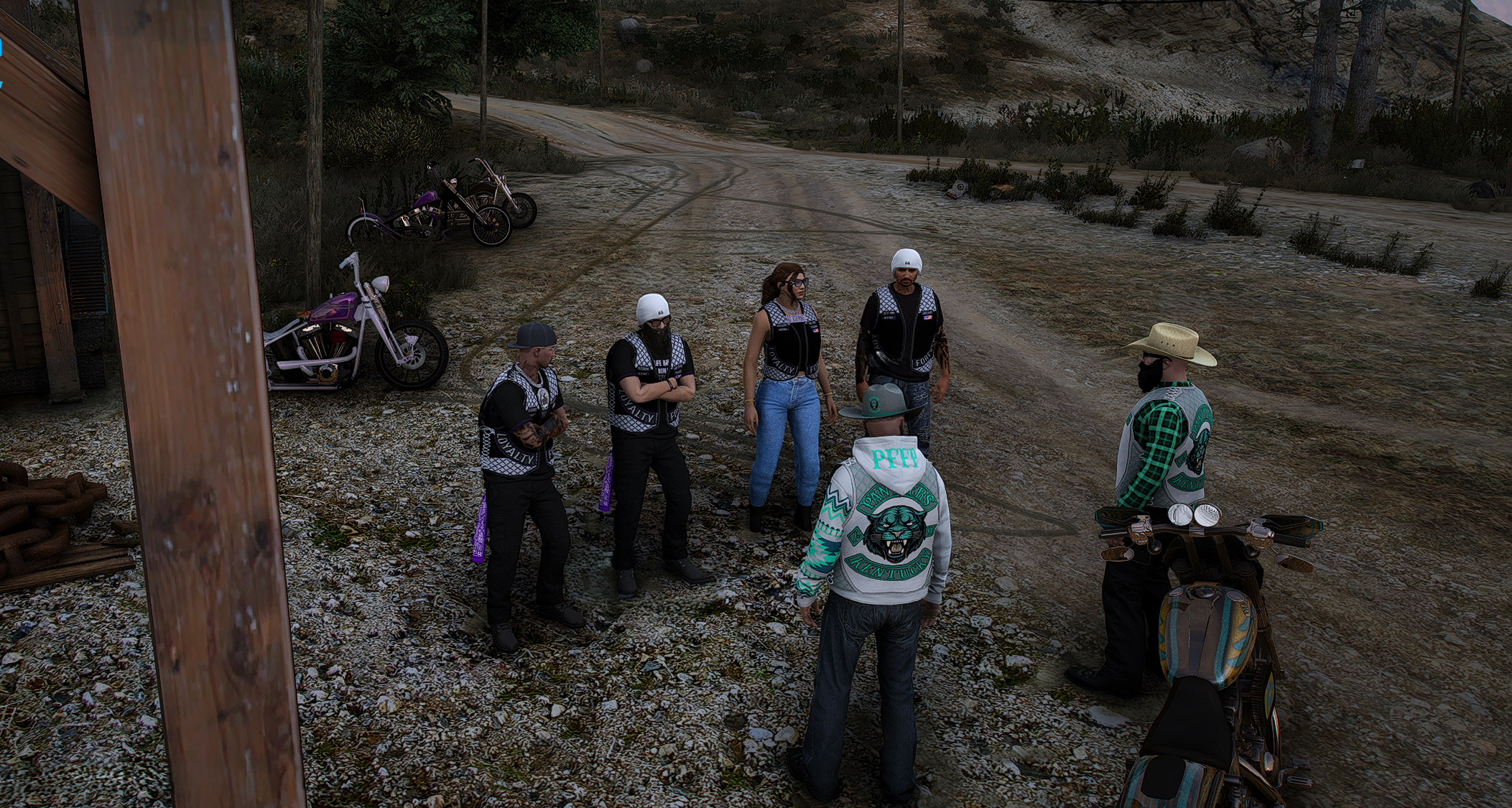 Group of six people, some wearing motorcycle gear and vests, standing on a dirt road near motorcycles with bikes parked nearby, in a rural outdoor setting.
