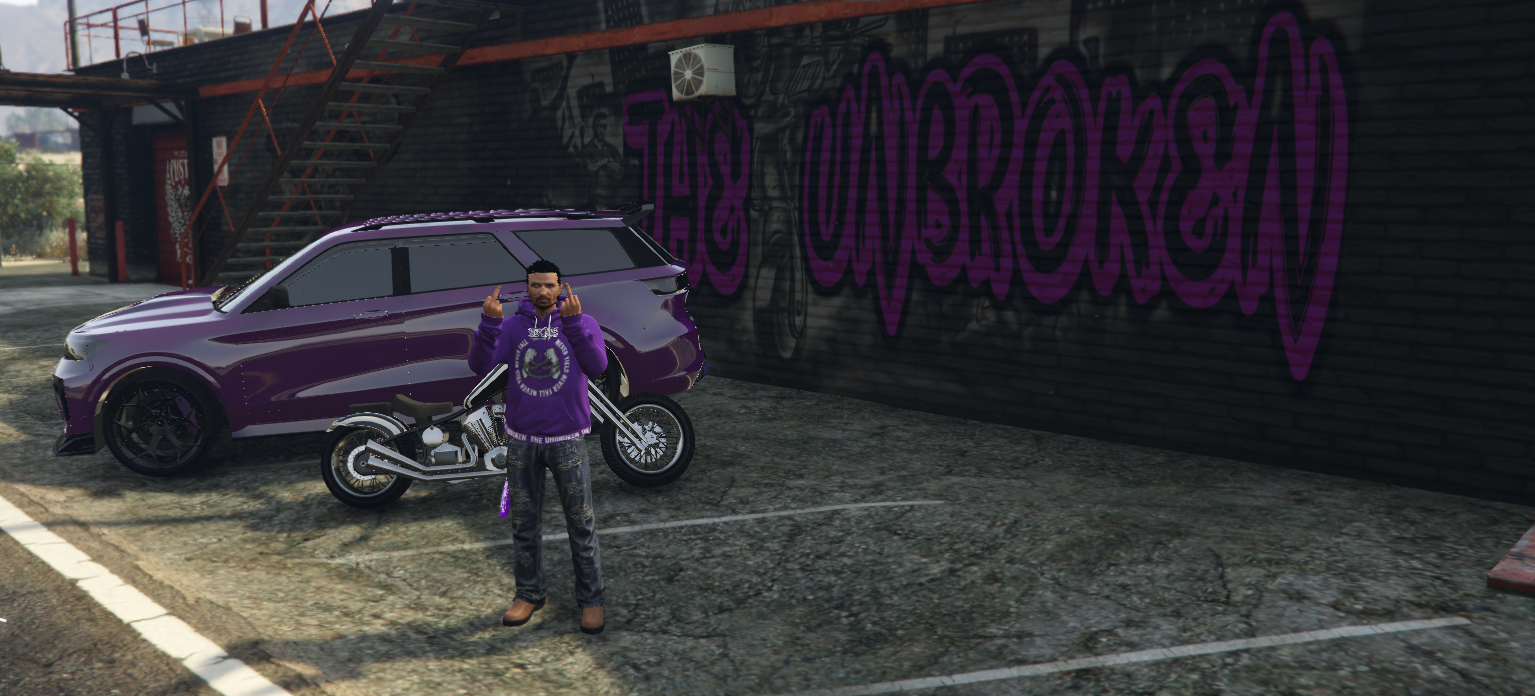 A man standing in front of a purple car and a motorcycle in a parking lot, with a graffiti wall in the background that reads '& TNumberK' in purple lettering.