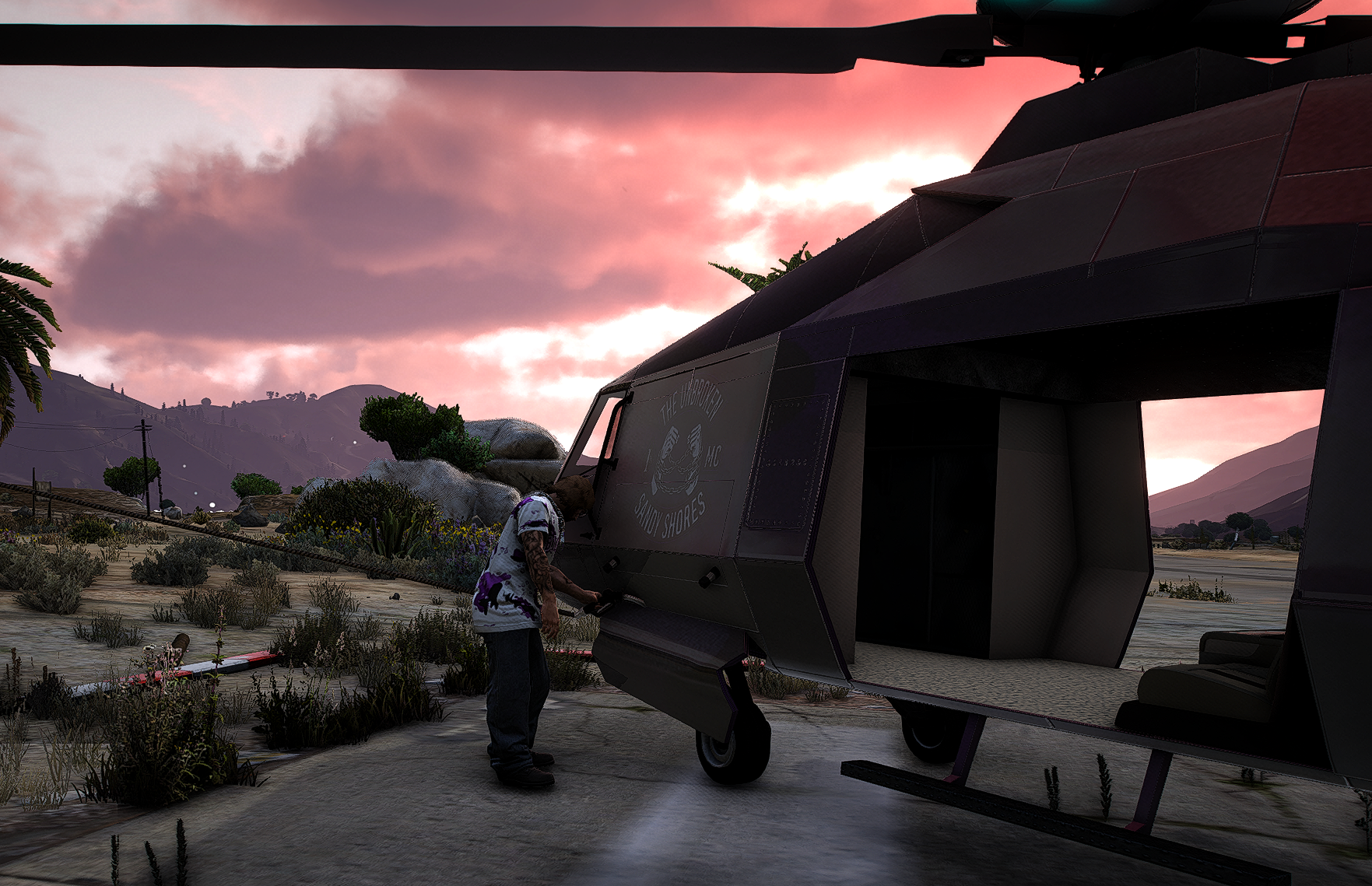 Person refueling a helicopter on a desert landscape during sunset, with mountains and vegetation in the background.