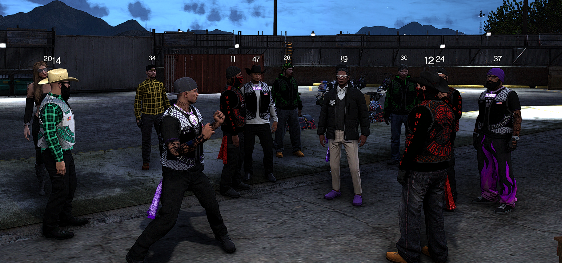 Group of people standing in a parking lot at dusk, some wearing biker vests and bandanas, with mountains in the background.