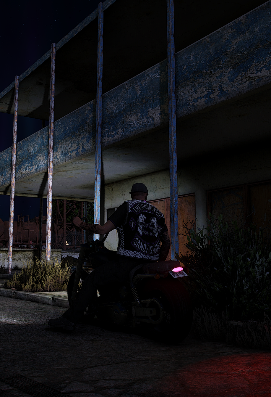 A person wearing a vest with a skull patch and the words 'The Broken' and 'Sandy Shores' on it, sitting on a motorcycle at night near a weathered building with metal bars and plants.