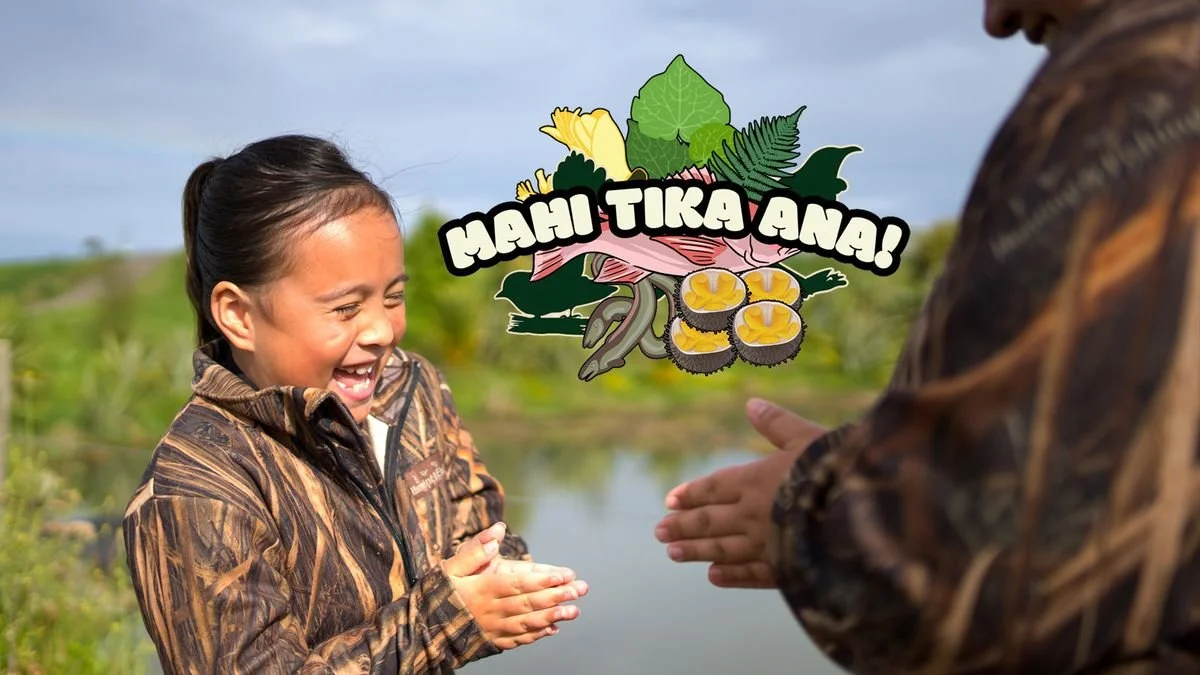 A young girl in camouflage clothing laughing as they enthusiastically greet each other by saluting in an outdoor setting near a body of water, with a cartoon graphic of seafood and the text "MAHI TIKA AN!" overlayed.