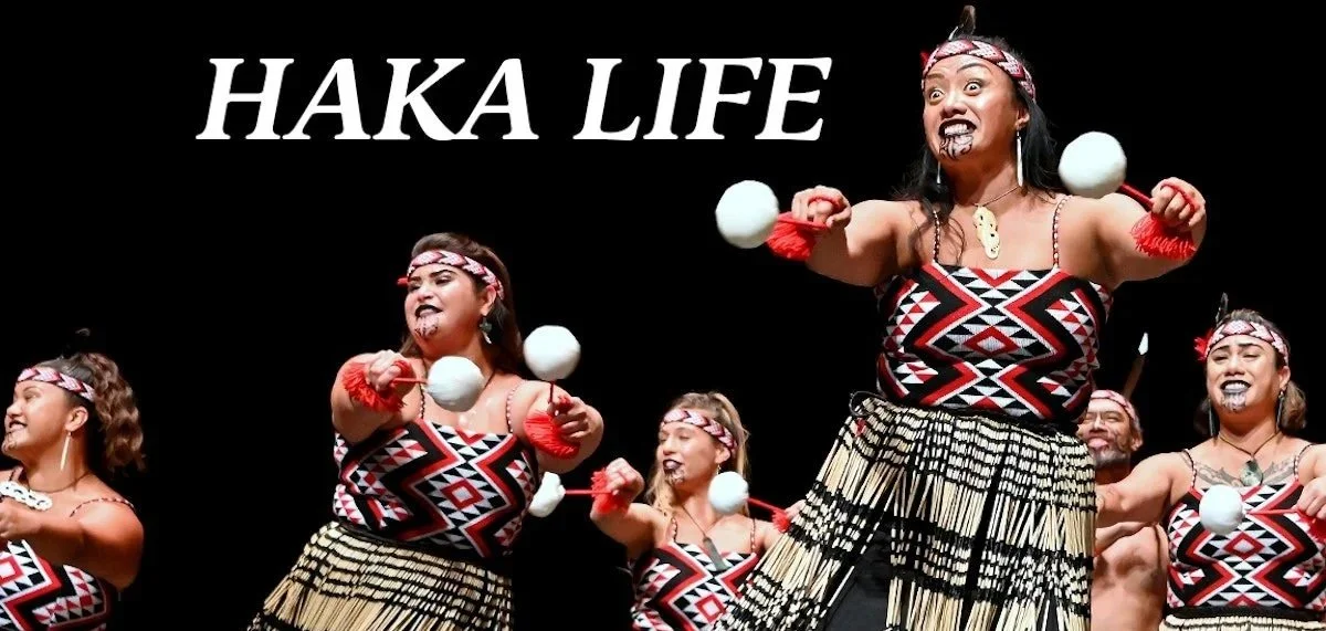 A group of performers in traditional Polynesian attire dancing and playing with white balls on red sticks, with face paint and headbands, on stage with the words 'HAKA LIFE' above them.