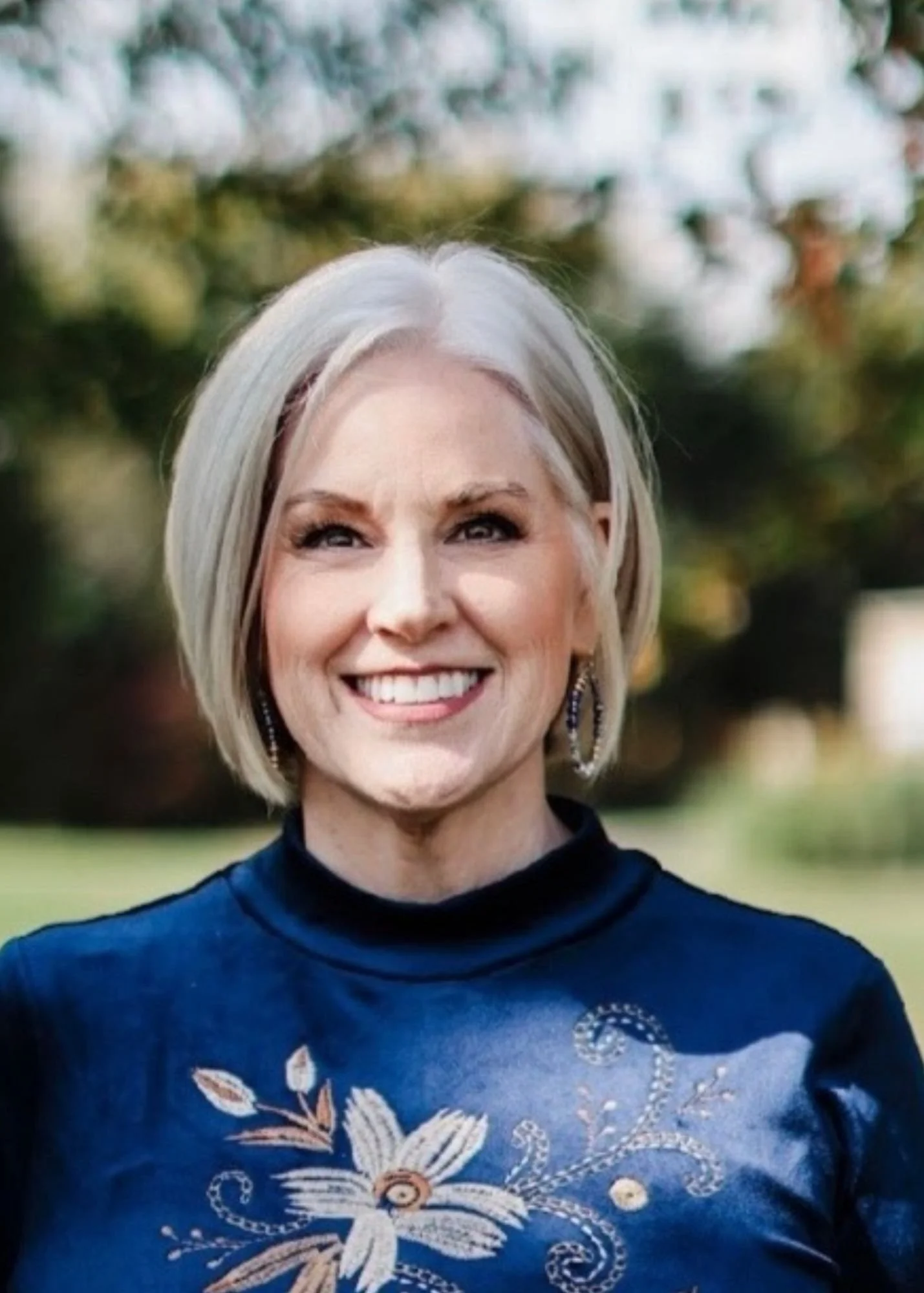 Front view of a woman with short blonde hair, smiling, wearing a navy blue top with embroidered floral and swirl designs, and earrings, outdoors with blurred greenery and trees in the background.