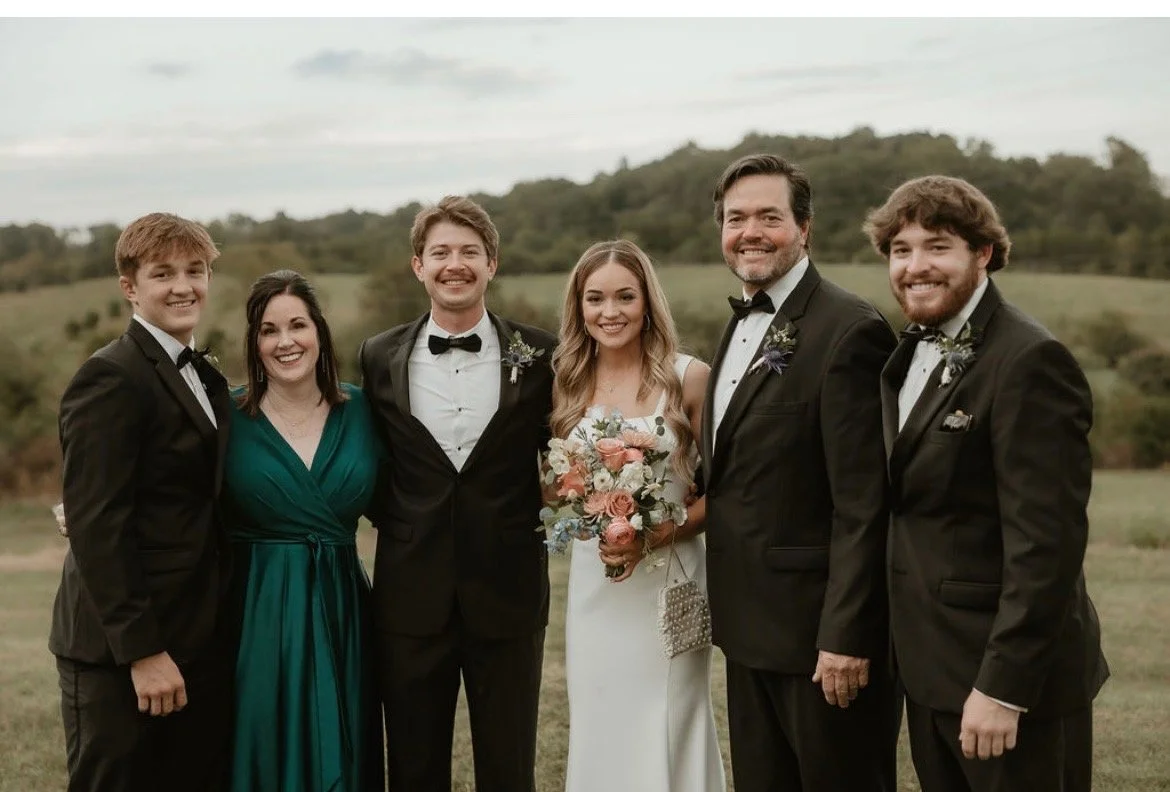 Group of six people in formal attire, smiling outdoors, during a wedding celebration with a scenic landscape background.