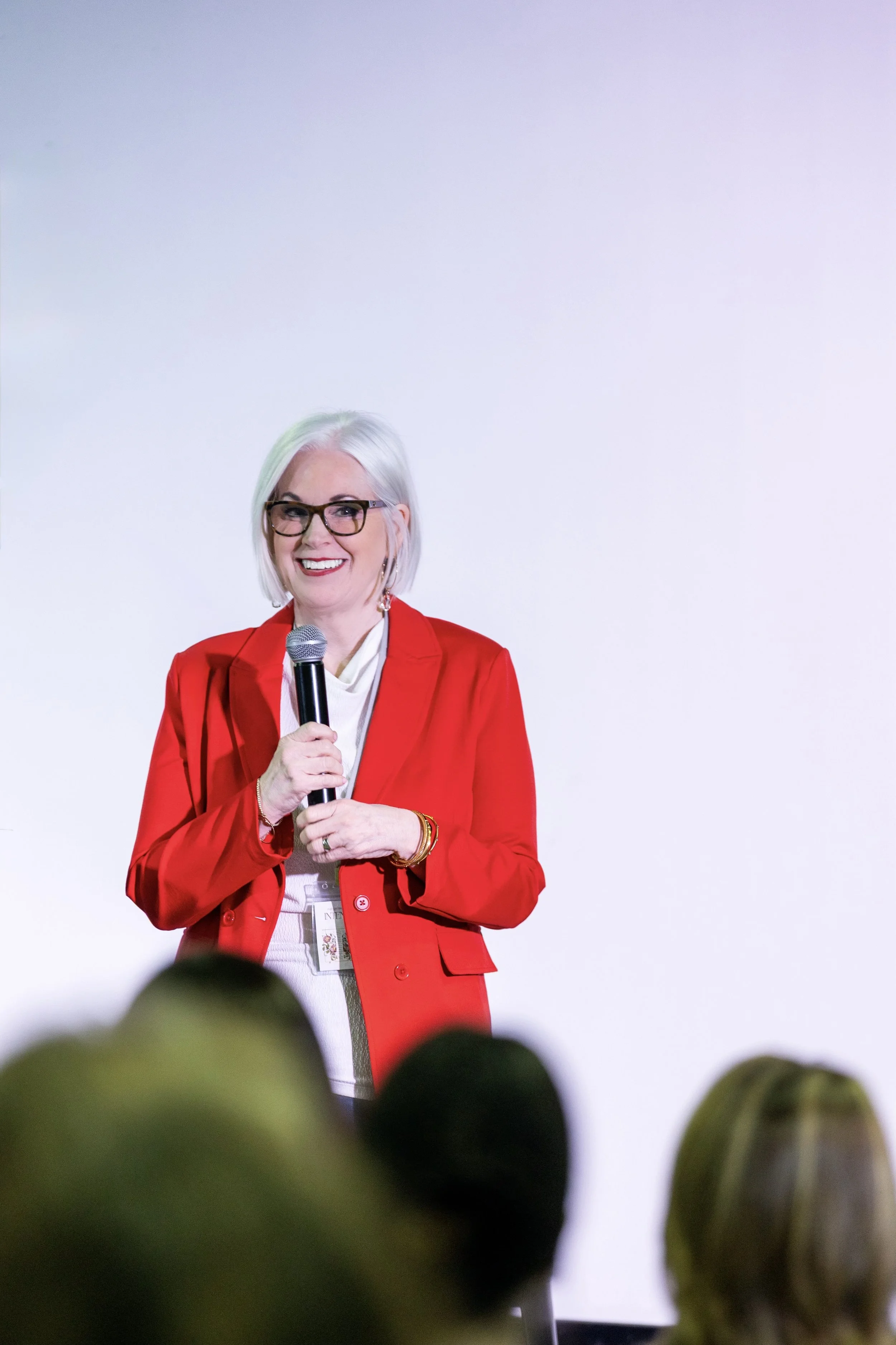 A woman with white hair, glasses, and a red blazer standing on stage holding a microphone, smiling, with an audience in front of her.
