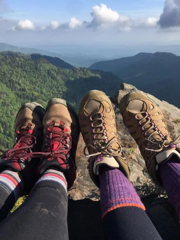 Two pairs of hiking boots on a rocky ledge overlooking green forested mountains and a blue sky with clouds.
