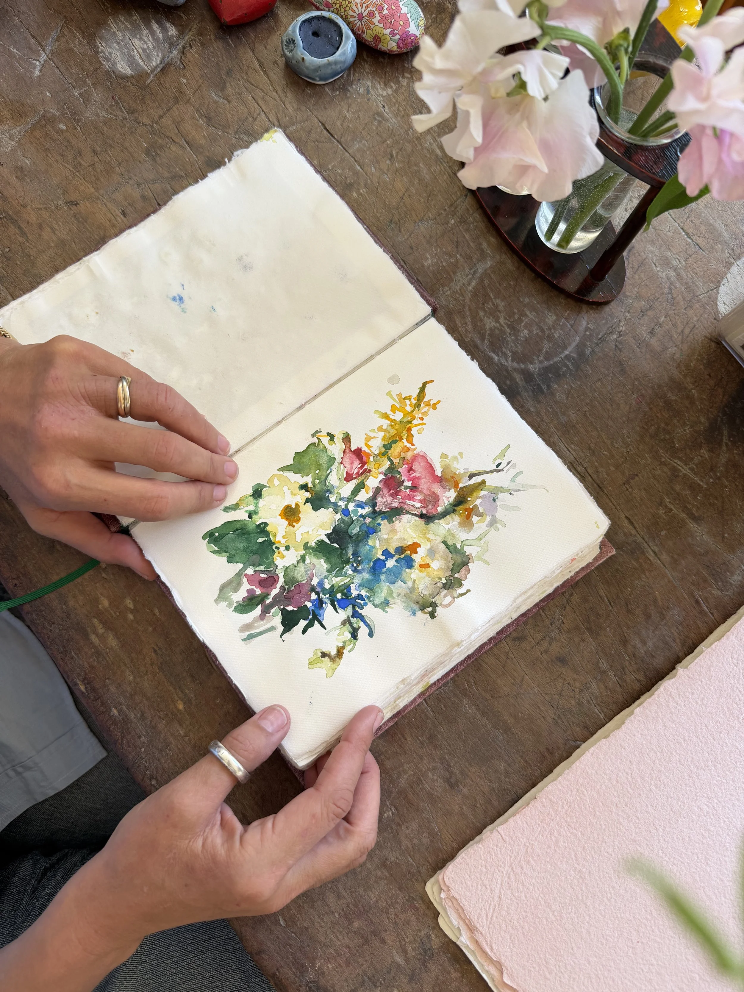 A person is holding an open sketchbook with a watercolor painting of a colorful bouquet of flowers on a wooden table. There are pink flowers in a glass vase, some small bowls, and a pink textured paper on the table.