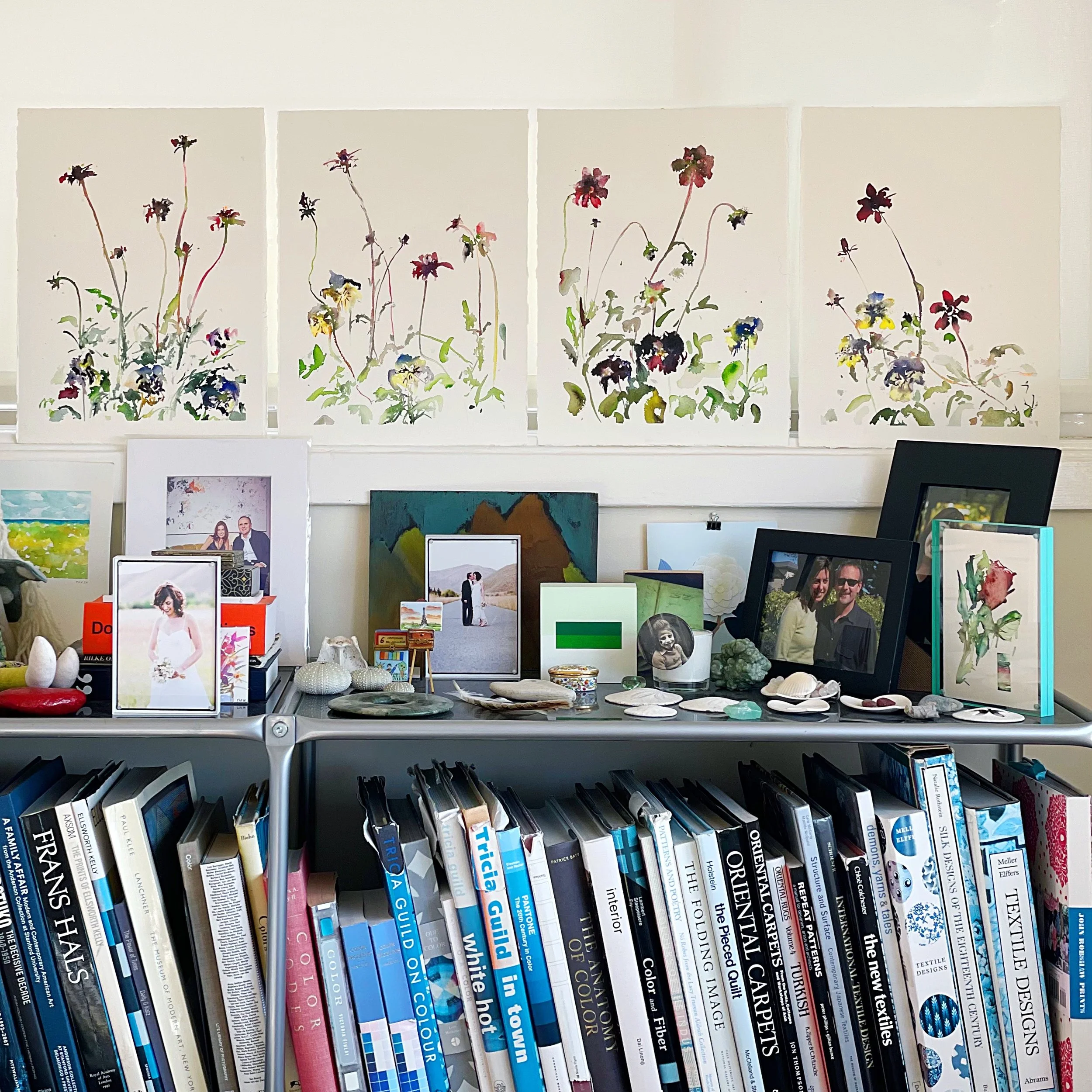 A shelf filled with framed photographs, books, shells, and decorative objects in front of four watercolor paintings of flowers on a white wall.