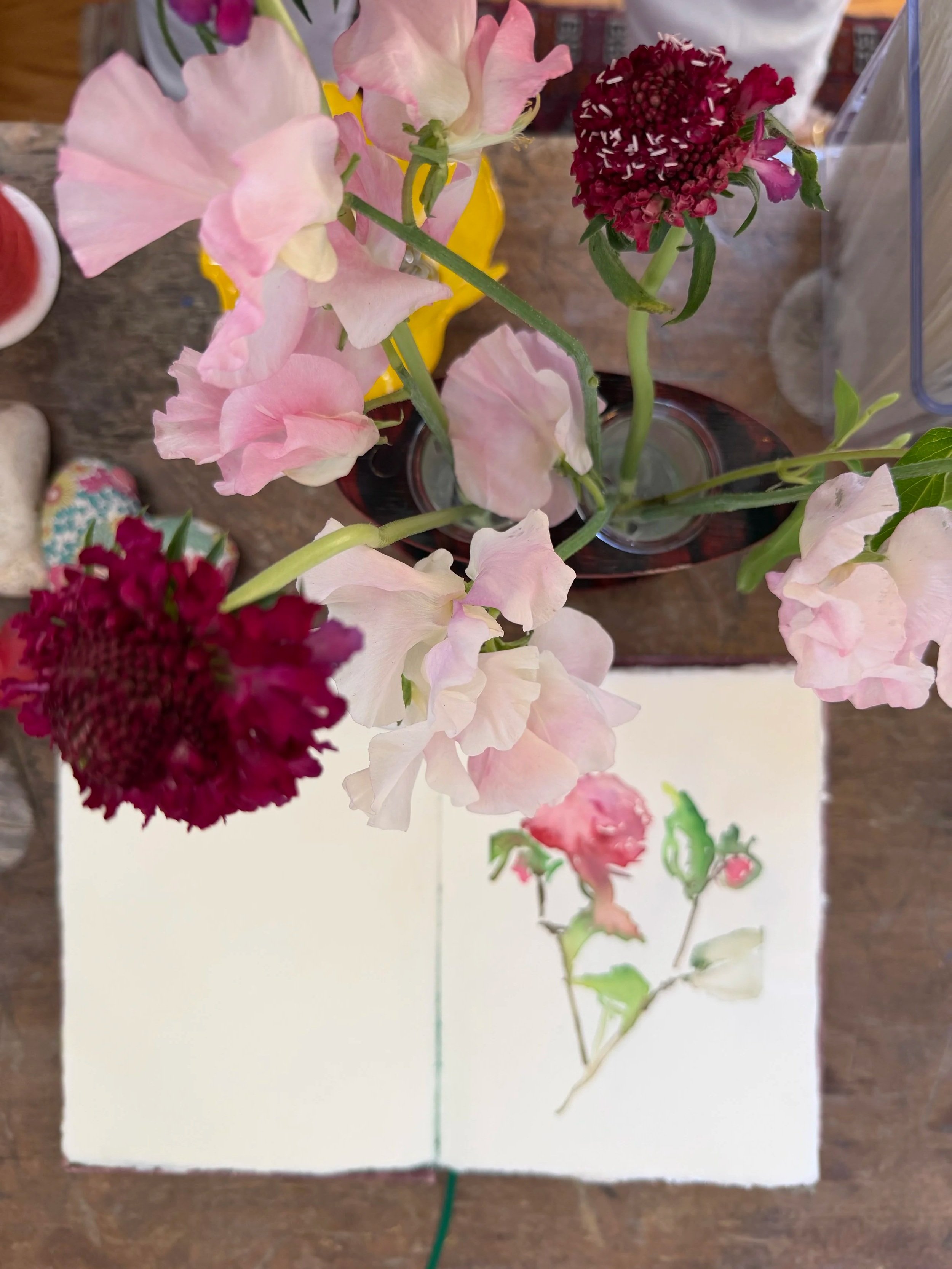 A bouquet of pink and deep red flowers on a wooden table with an open watercolor sketchbook in the background.