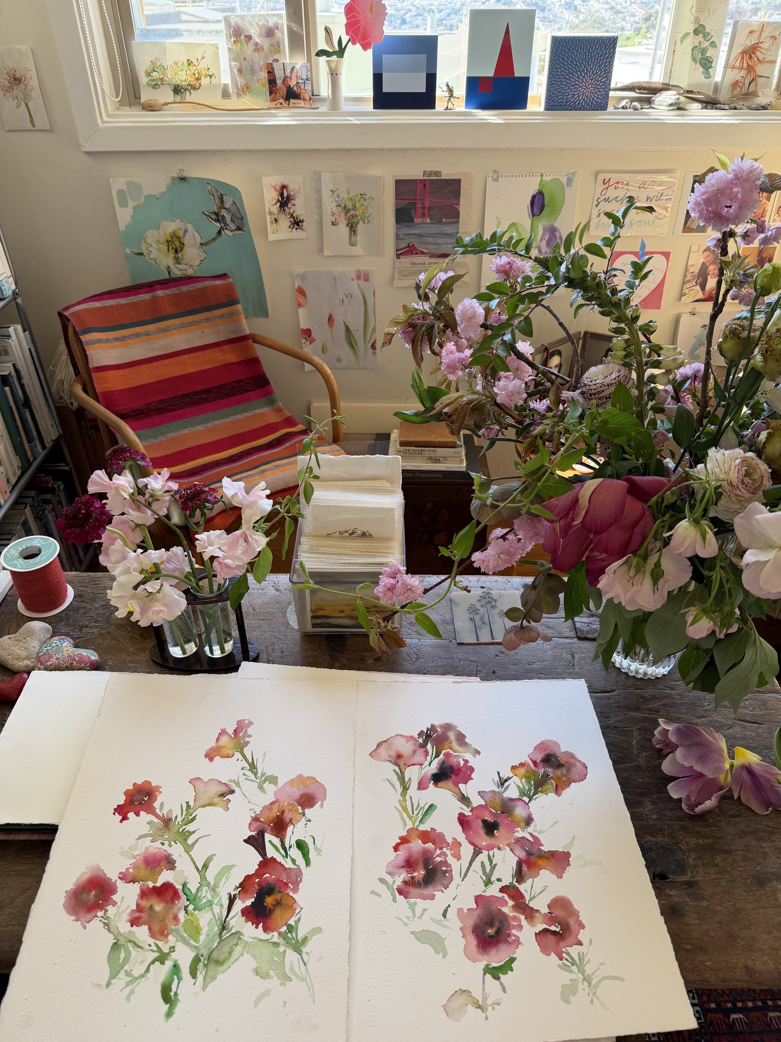 A table with watercolor paintings of pink and red flowers, a glass vase with pink flowers, and various art supplies. The background shows a window ledge with decorative items and a wall with attached artwork, including a painting of a hare and severa
