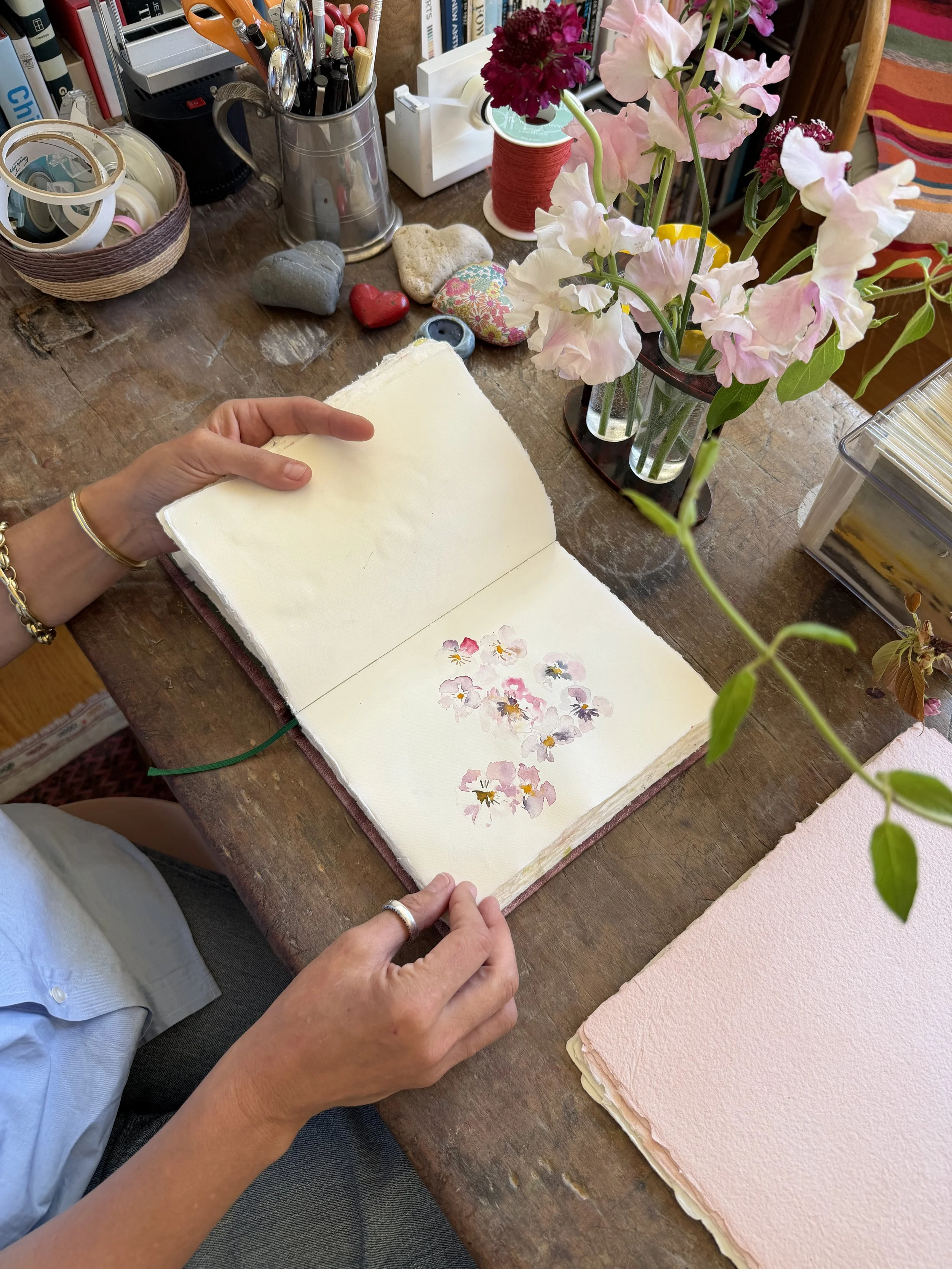 A person is flipping through an open sketchbook with watercolor floral paintings inside. The sketchbook is on a wooden table along with a bouquet of pink and purple flowers, art supplies, decorative stones, and craft materials.