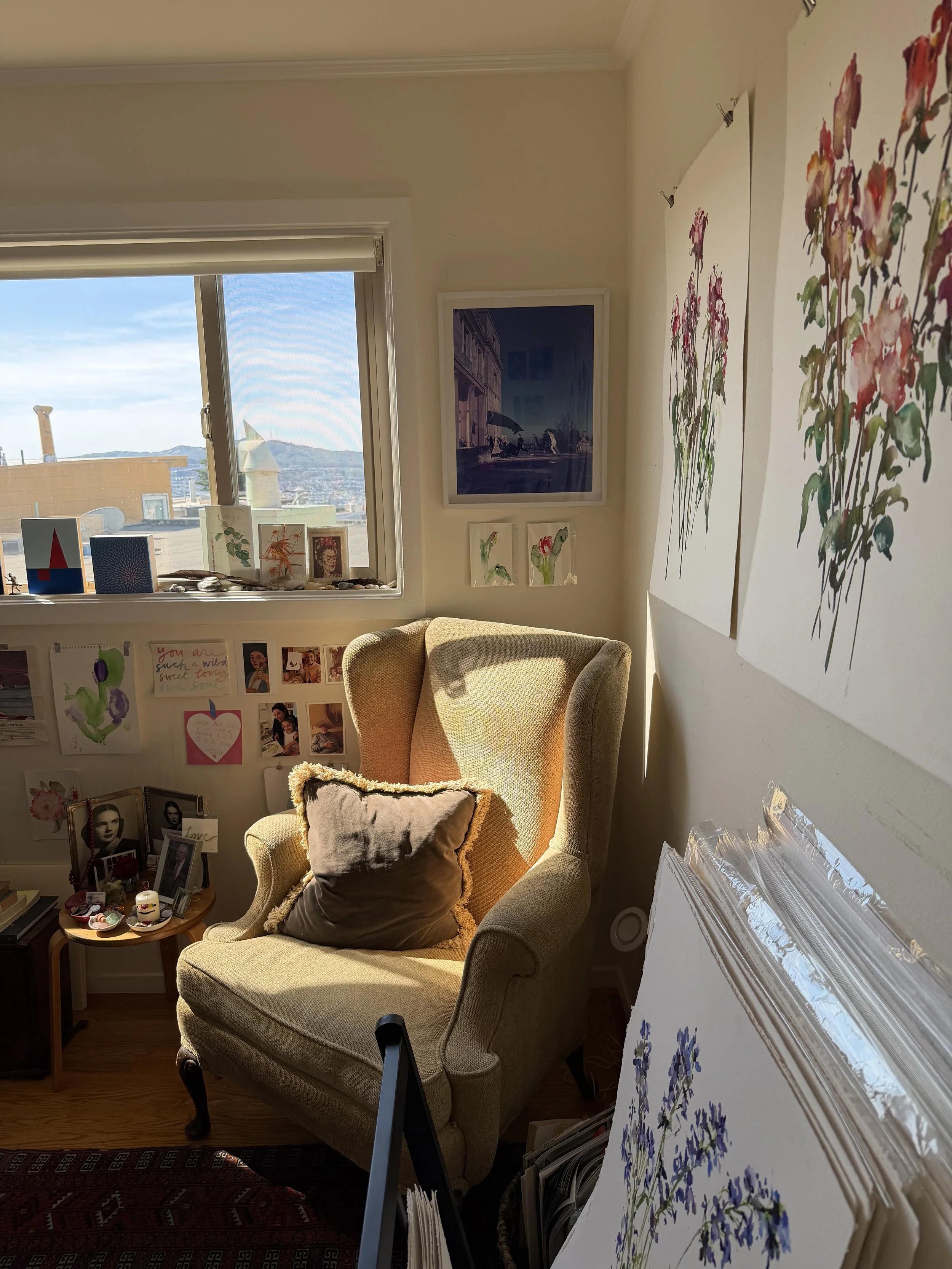 A cozy corner of a room with a yellow armchair, decorated with a brown cushion. To the left, a small side table with photographs and decorative items. The room has artwork on the walls, including floral paintings, and a window with a view of a citysc
