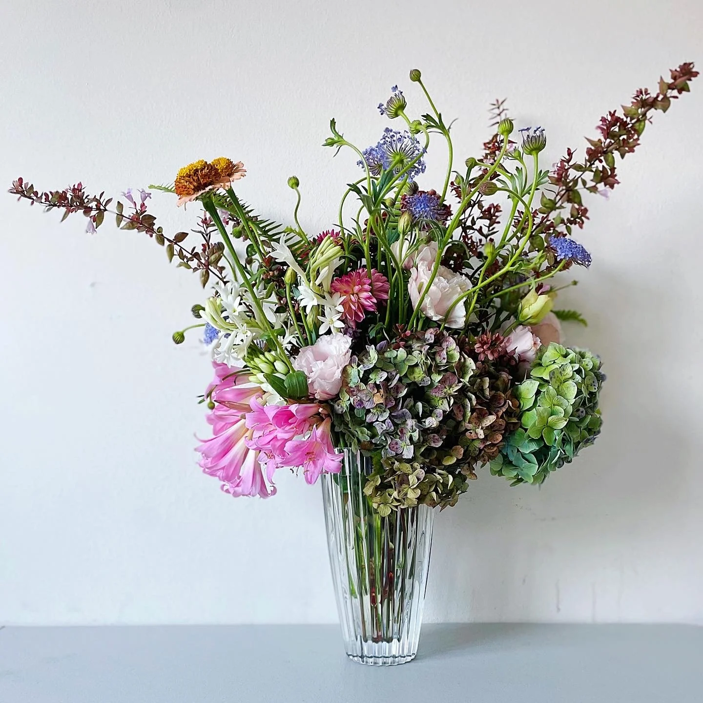 A tall, clear glass vase filled with a colorful bouquet of various flowers including pink lilies, purple hydrangeas, white star-shaped flowers, and small purple and green accents, set against a plain white background.