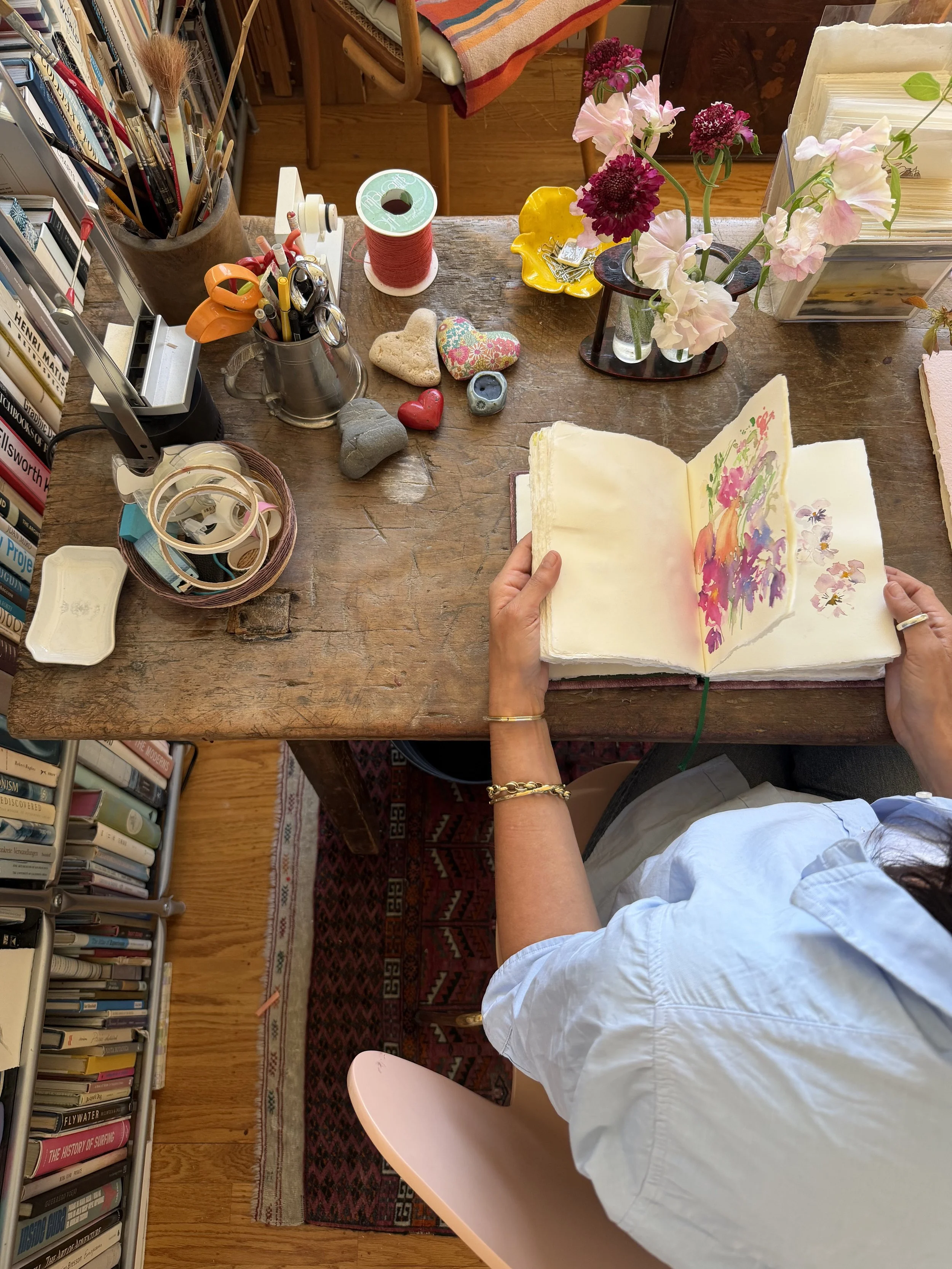 A person holding an open sketchbook with a floral watercolor painting, sitting at a cluttered wooden desk with art supplies, a vase of pink and purple flowers, and books nearby.