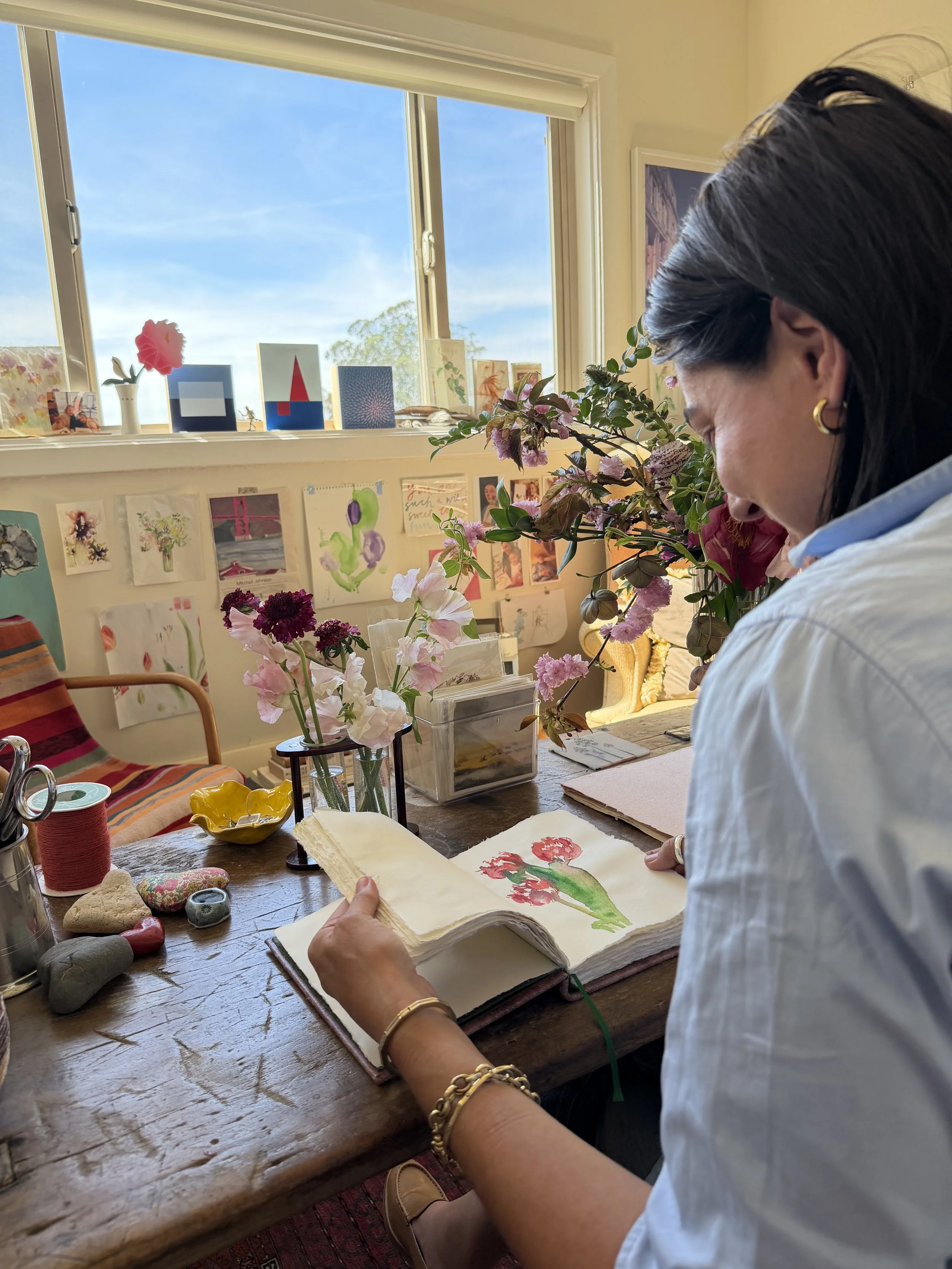 Woman sitting at a wooden table, looking at a watercolor painting of a pink flower in a sketchbook, with a bright window behind her and various art supplies, flowers, and artwork on the table and window sill.