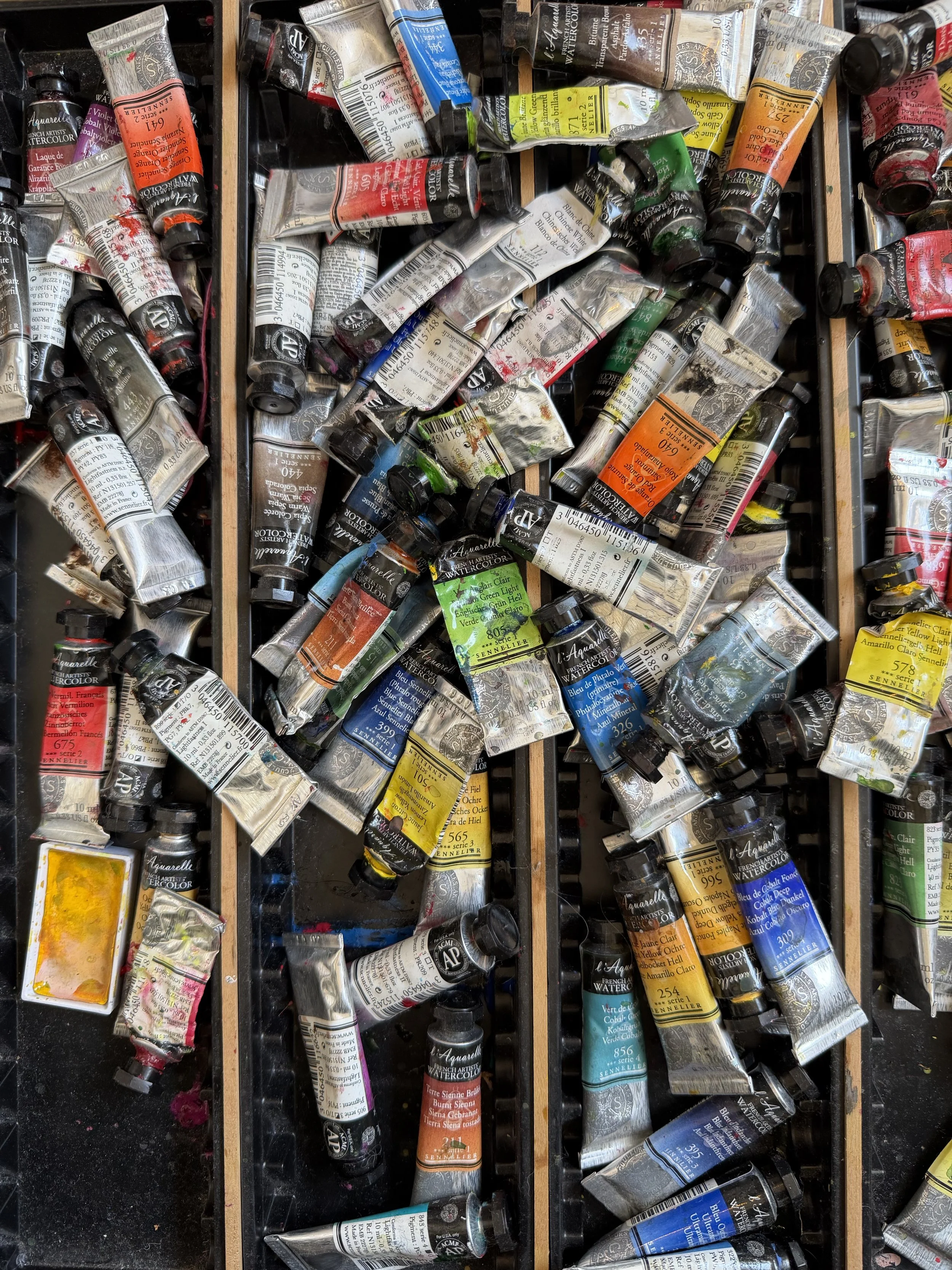 A messy collection of watercolor paint tubes in various colors scattered in compartments.