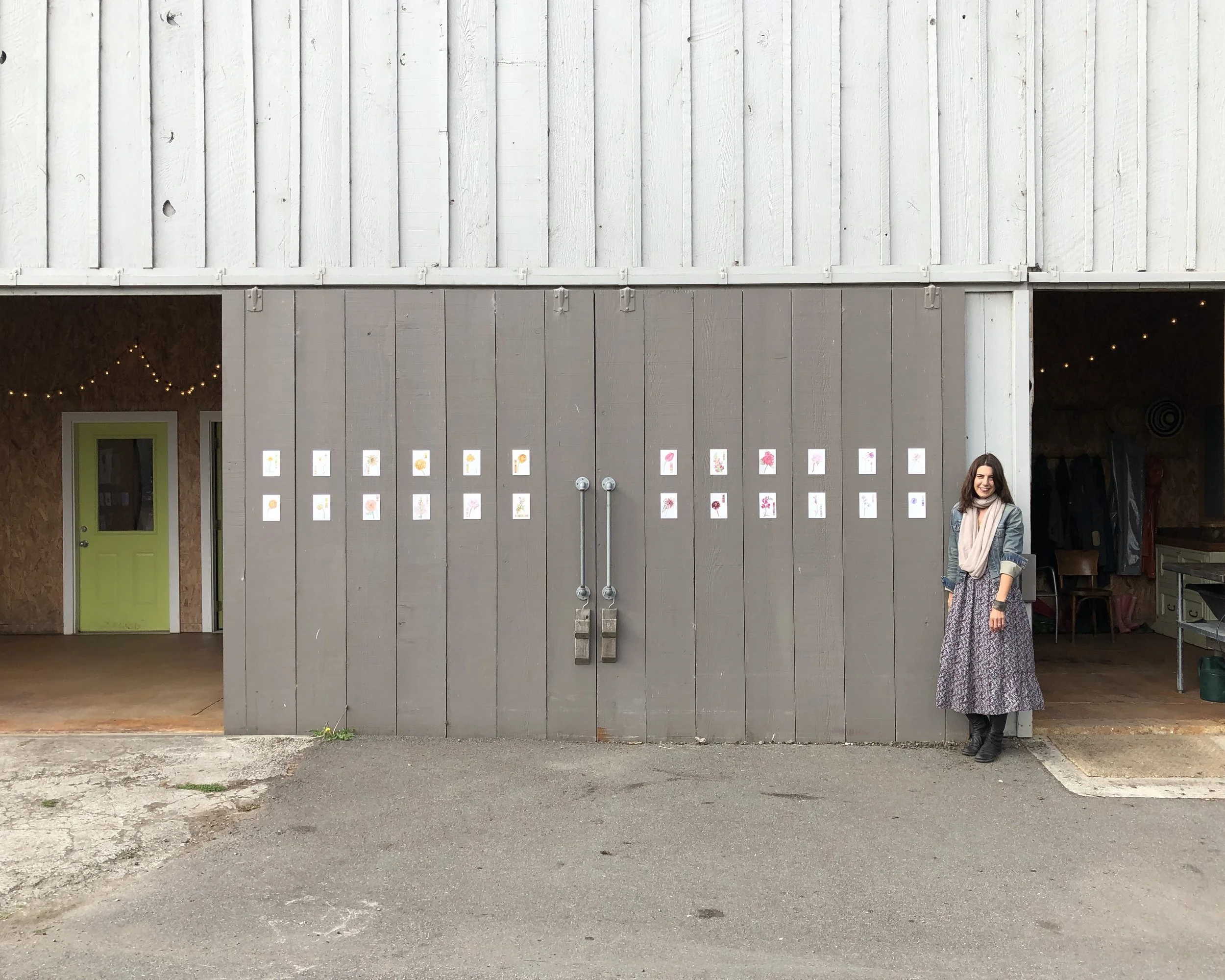 Woman standing outside a barn with artwork displayed on the door, featuring various colorful floral illustrations.