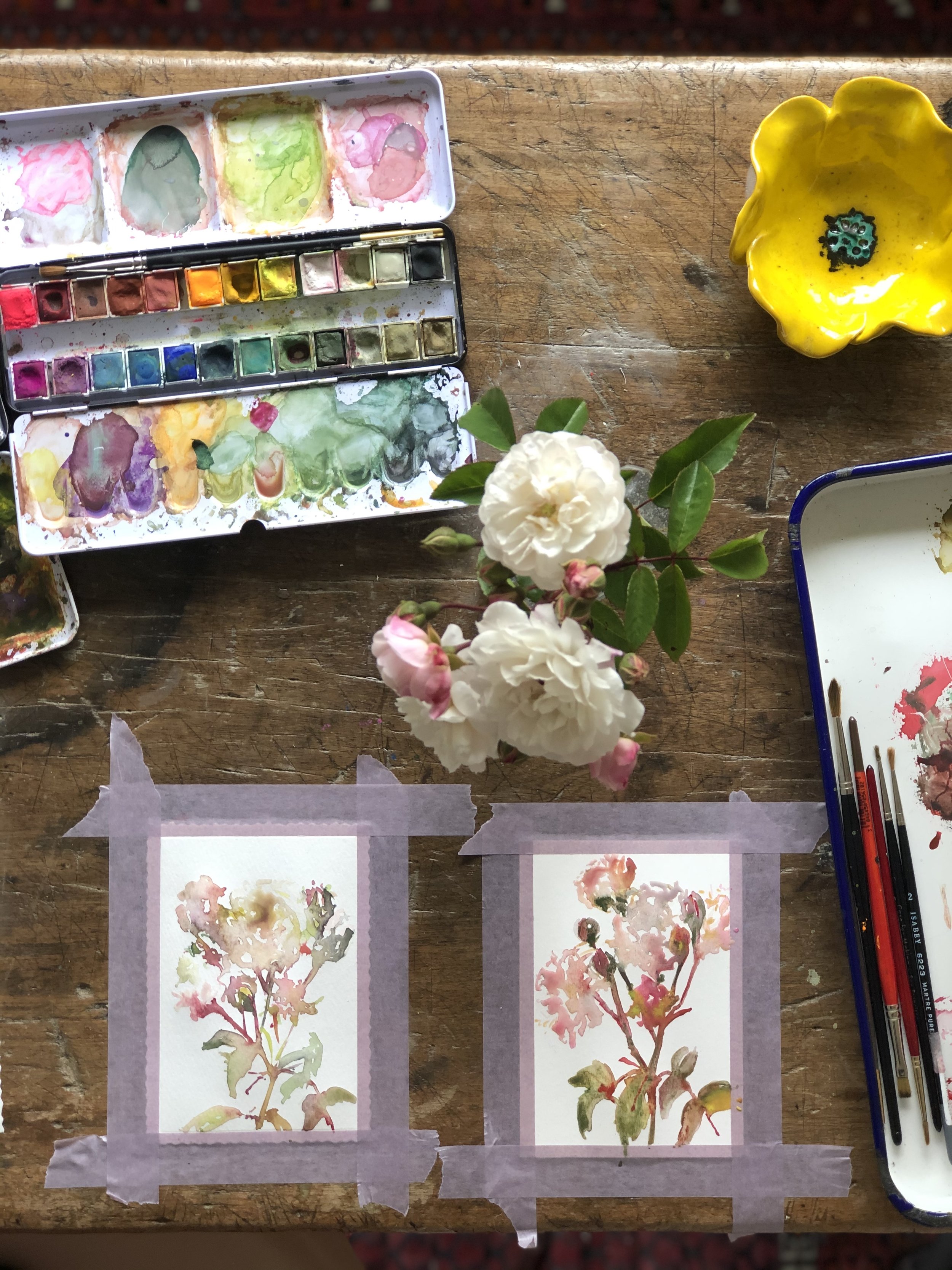 Watercolor painting supplies on a wooden table, including a watercolor palette, brushes, and two small watercolor paintings of pink flowers taped to the table. There is a yellow ceramic bowl and a white flower with pink buds in the center.
