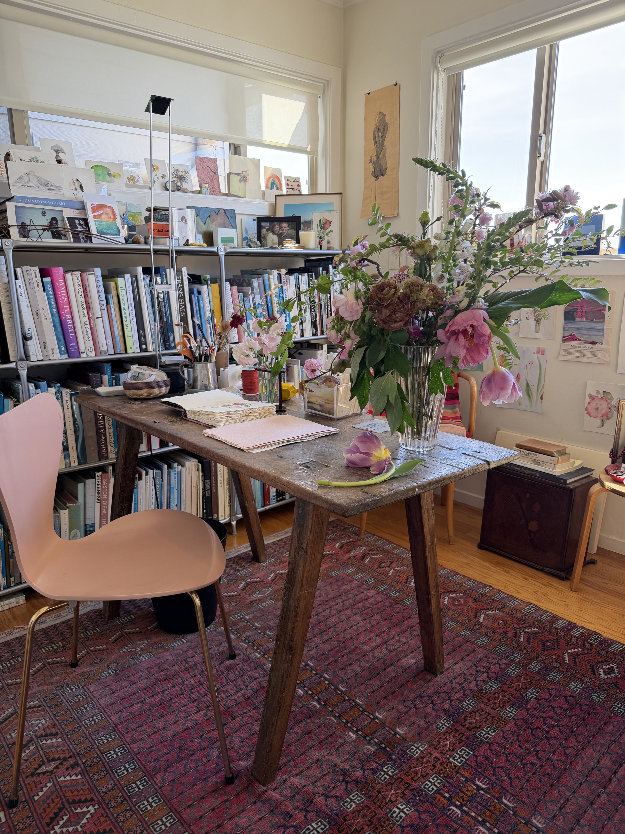 A cozy room with a wooden table holding a large bouquet of pink and purple flowers in a glass vase. There's a pink chair beside the table, a bookshelf filled with books, and a window with a view outside. The room has various artworks and photos on th