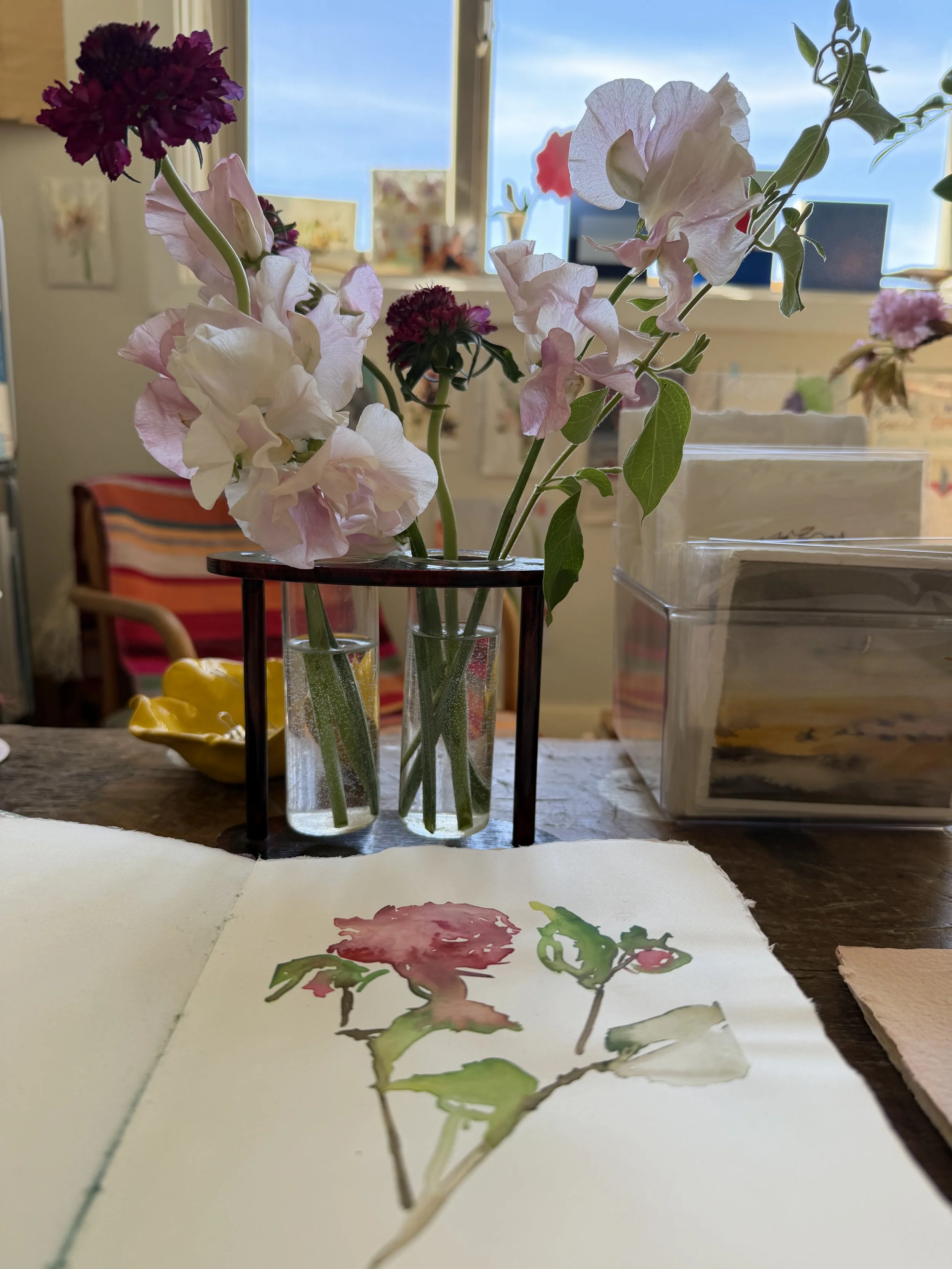 A vase with pink and purple flowers on a table, with watercolor sketches of the flowers on paper in the foreground.