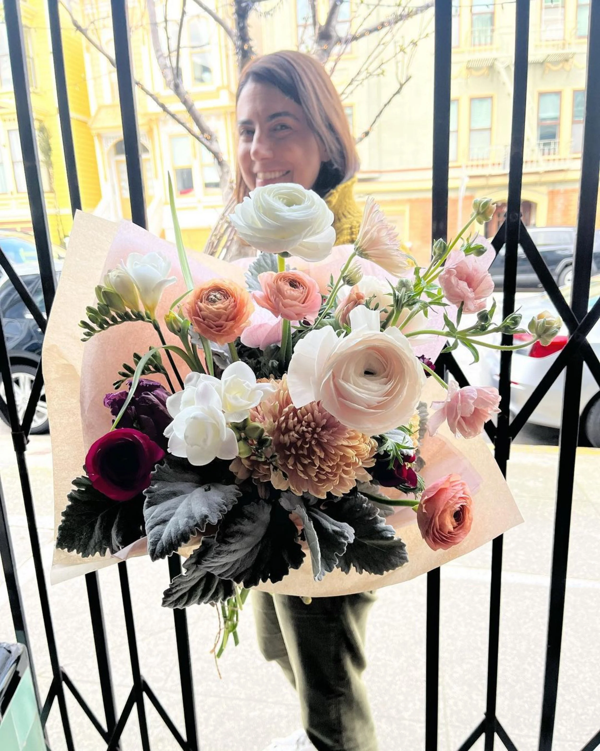 Woman holding a bouquet of pink, white, and purple flowers outside a building with colorful architecture, seen through a black metal gate